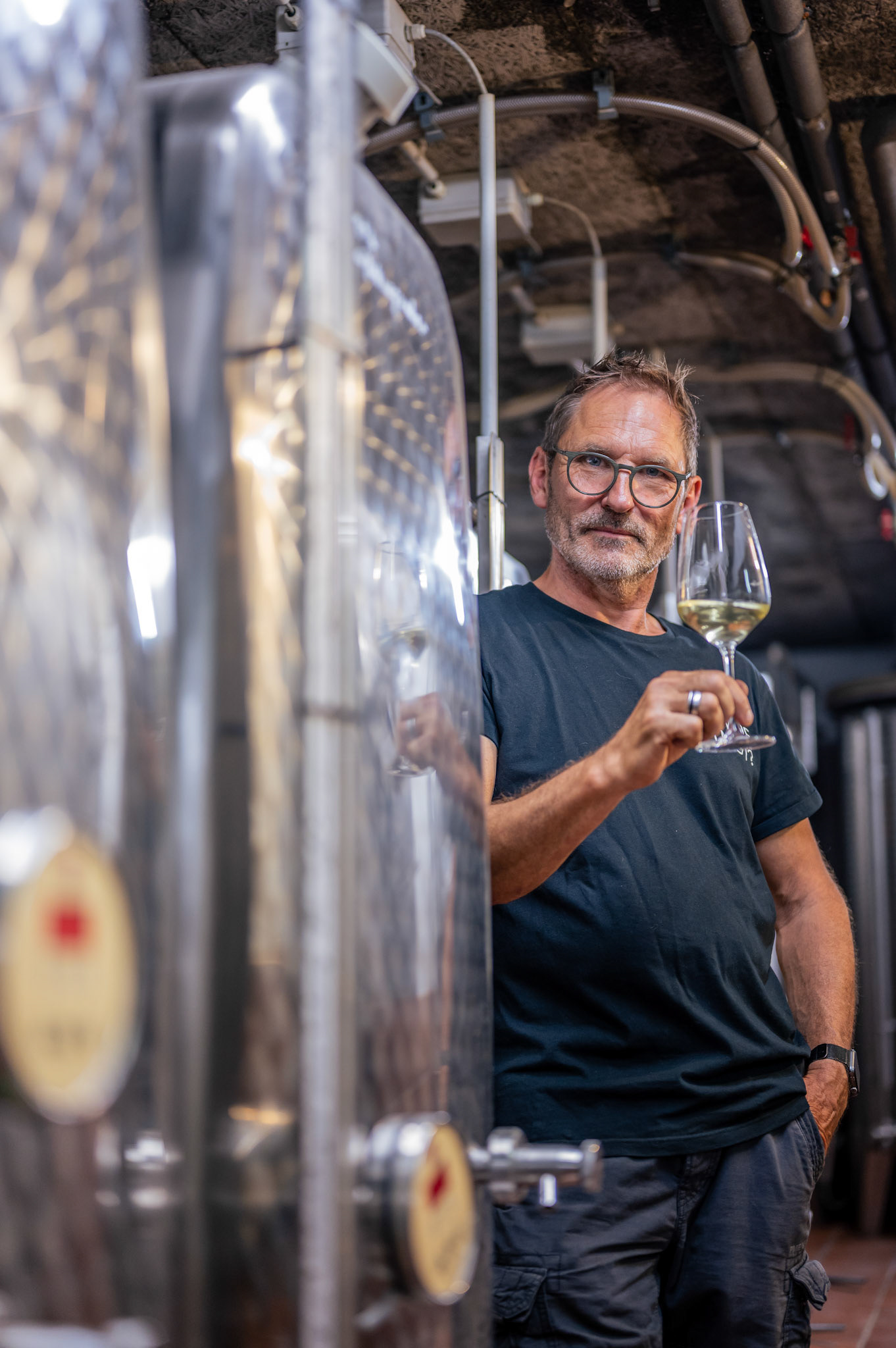 Mann steht mit einem Glas Wein im Weinkeller an Weintanks