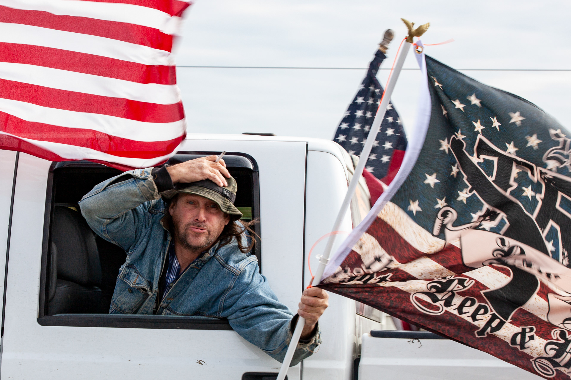 Trump Parade participant waves a flag out of the window on I-270 while holding onto his hat in Columbus, Ohio on Oct. 3, 2020. Trump supporters showed up in large numbers on I-270 to form a parade line on the highway encircling Columbus, Ohio, from 10am to noon to show support for President Donald Trump during election season.