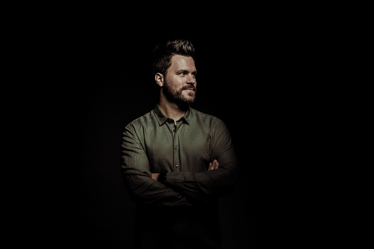 professionelle Studiofotografie Gabler, junger Mann in dunkelgrünem Hemd steht vor schwarzem Hintergrund mit verschränkten Armen und blickt zur Seite, nur der Oberkörper ist gezielt beleuchtet