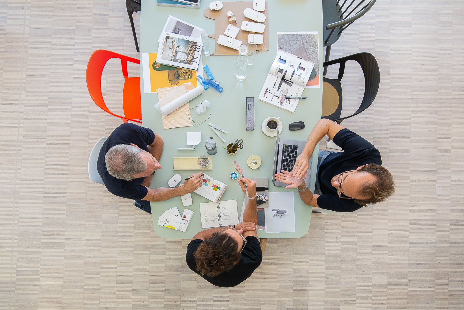 Interiorfotografie Christoph Gabler, Drei Designer sitzen an hellem Tisch mit bunten Stühlen und diskutieren in typischer Arbeitssituation, Vogelperspektive