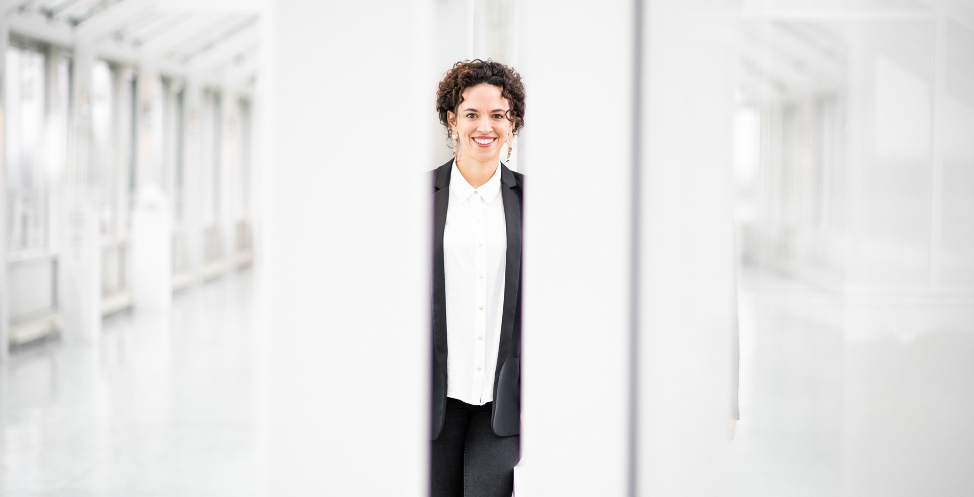 Businessfoto Regensburg, junge Frau in schwarzem Anzug mit weißem Hemd steht mittig in lichtdurchfluteter Location mit vielen Fenstern, fotografiert im Anschnitt eines Fensters mit Spiegelung und einer weißen Säule
