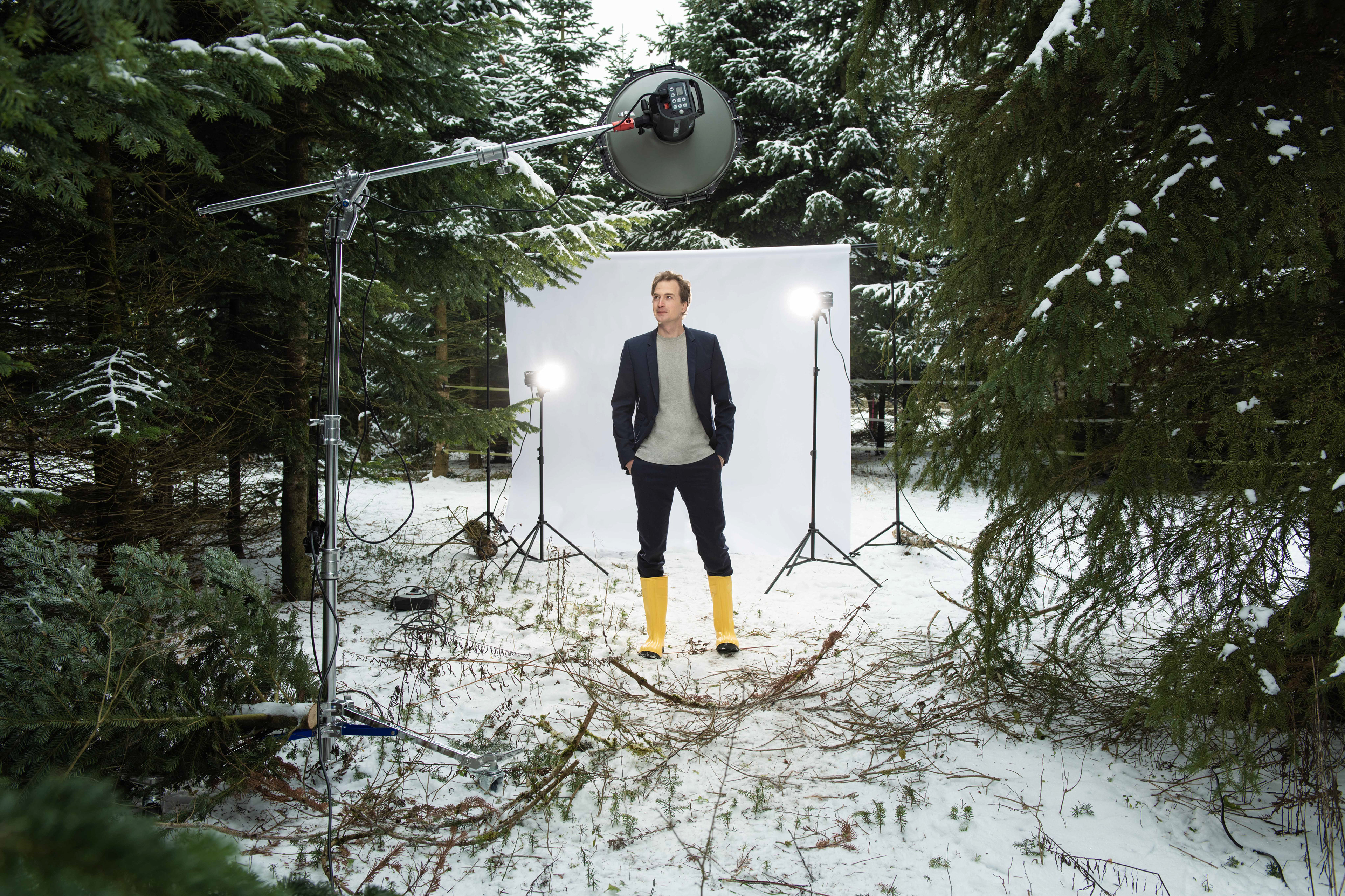 Fotograf Regensburg: Mann in schwarzem Sakko und Hose sowie gelben Gummistiefeln steht vor weißer Leinwand, die mit drei Fotoblitzen im Wald aufgebaut wurde