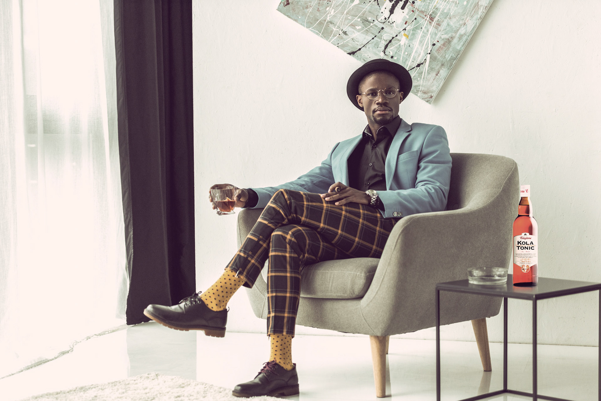 stylish african american man in hat, checkered pants and blue jacket with cigar and glass of cognac sitting in armchair in modern office