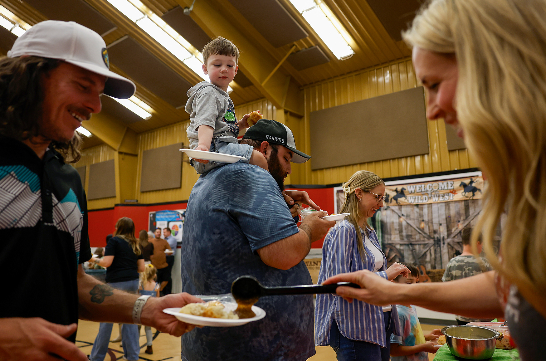 Milo Varvaro, 2, center, sits on John Varvaro’s shoulders as Principal Amanda Bruckerhoff serves food to Matt McDerman on March 19 at Two Mile Prairie Elementary School. John Varvaro tasted drop biscuits and crockpot strawberry jam made by the third graders. “They are really great,” John Varvaro said.