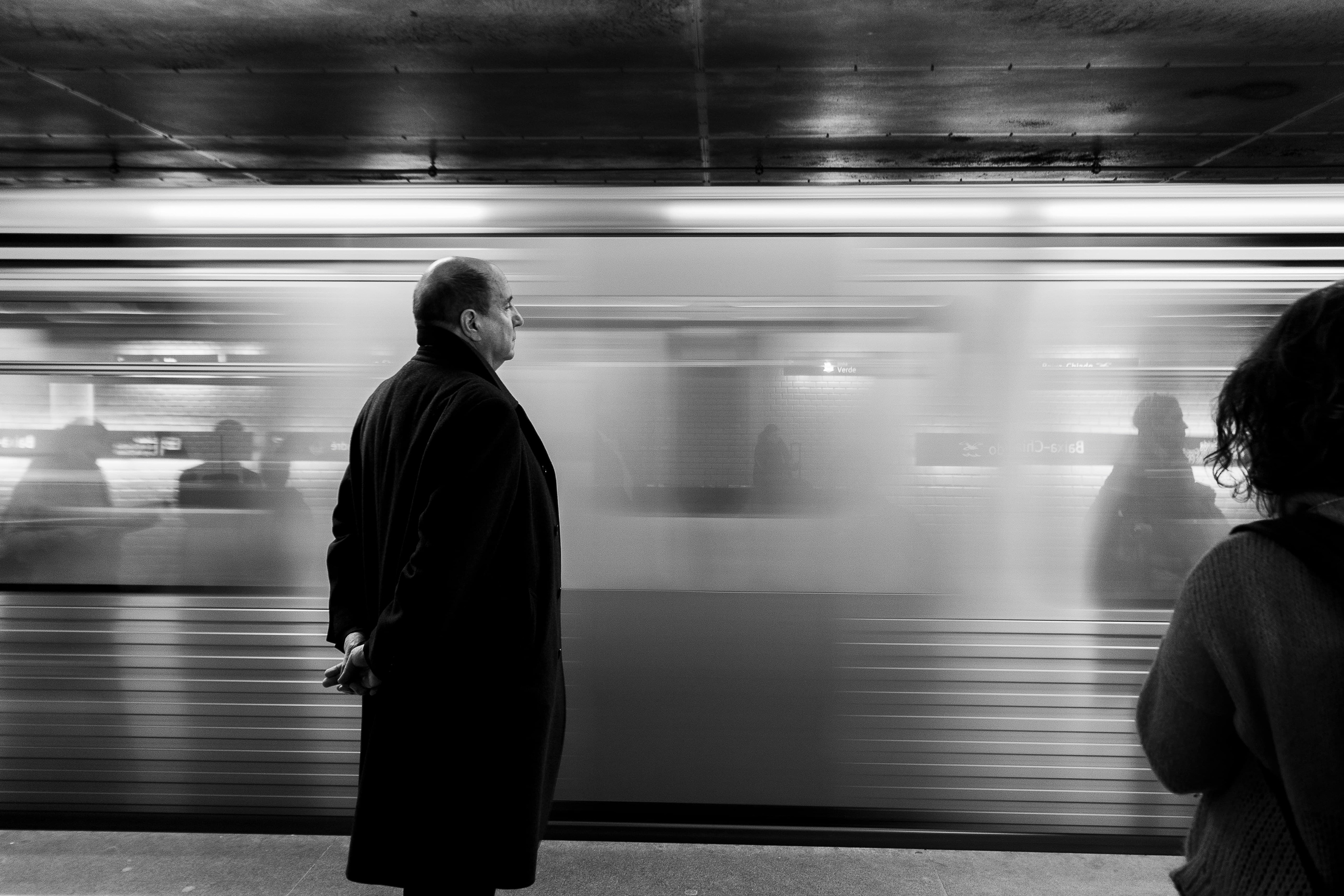 A man stands with his arms crossed behind his back as a subway train pulls in. The man is in sharp focus while the arriving subway train is blurred. Black and white photo.