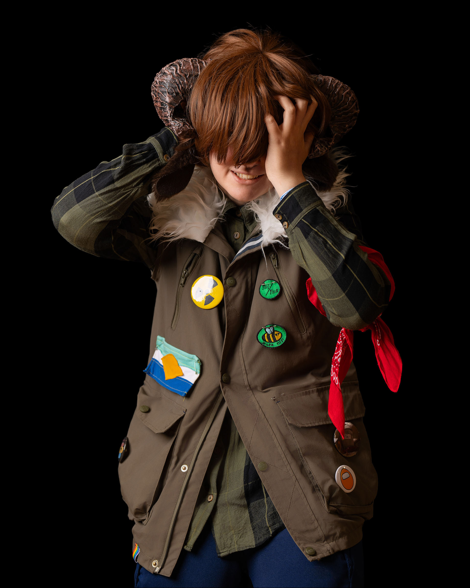 Studio cosplay image of King adjusting their horn headpiece, smiling with layered costume and unique character expression against a dark backdrop.