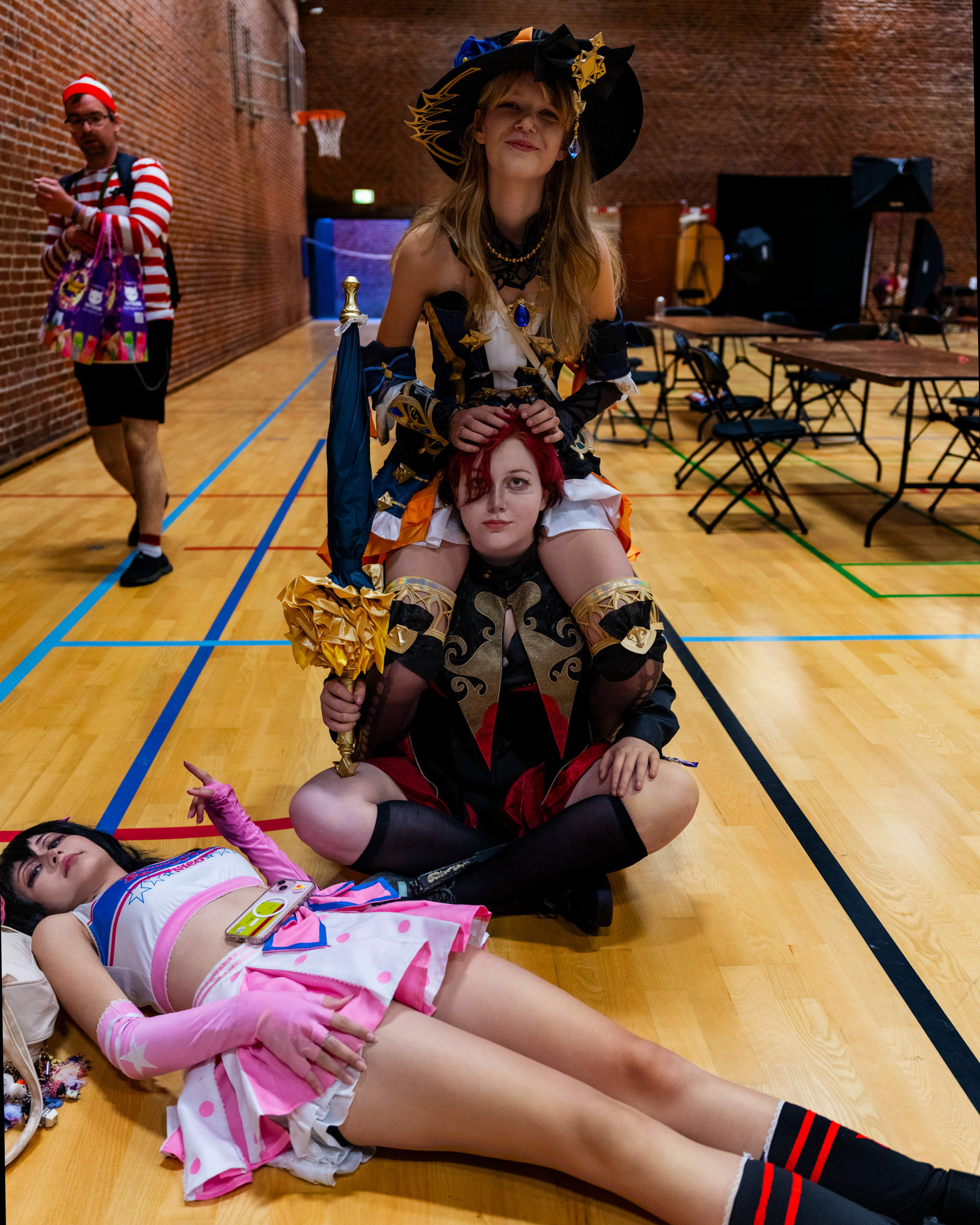 Cosplayer ruxqym sitting playfully on the shoulders of cosplayer zen.c0z, while cosplayer may_c0z lies on the floor in a cheerleader-inspired outfit inside a convention hall.