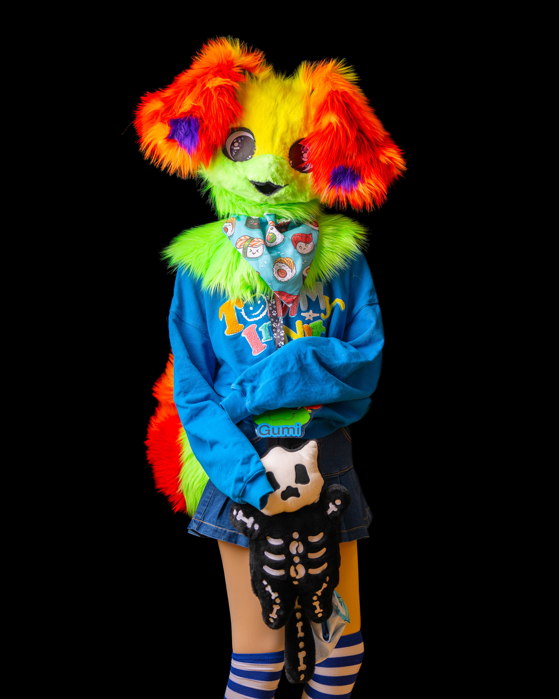 Furry cosplay character standing centered in studio wearing blue hoodie outfit and holding skeleton plush.