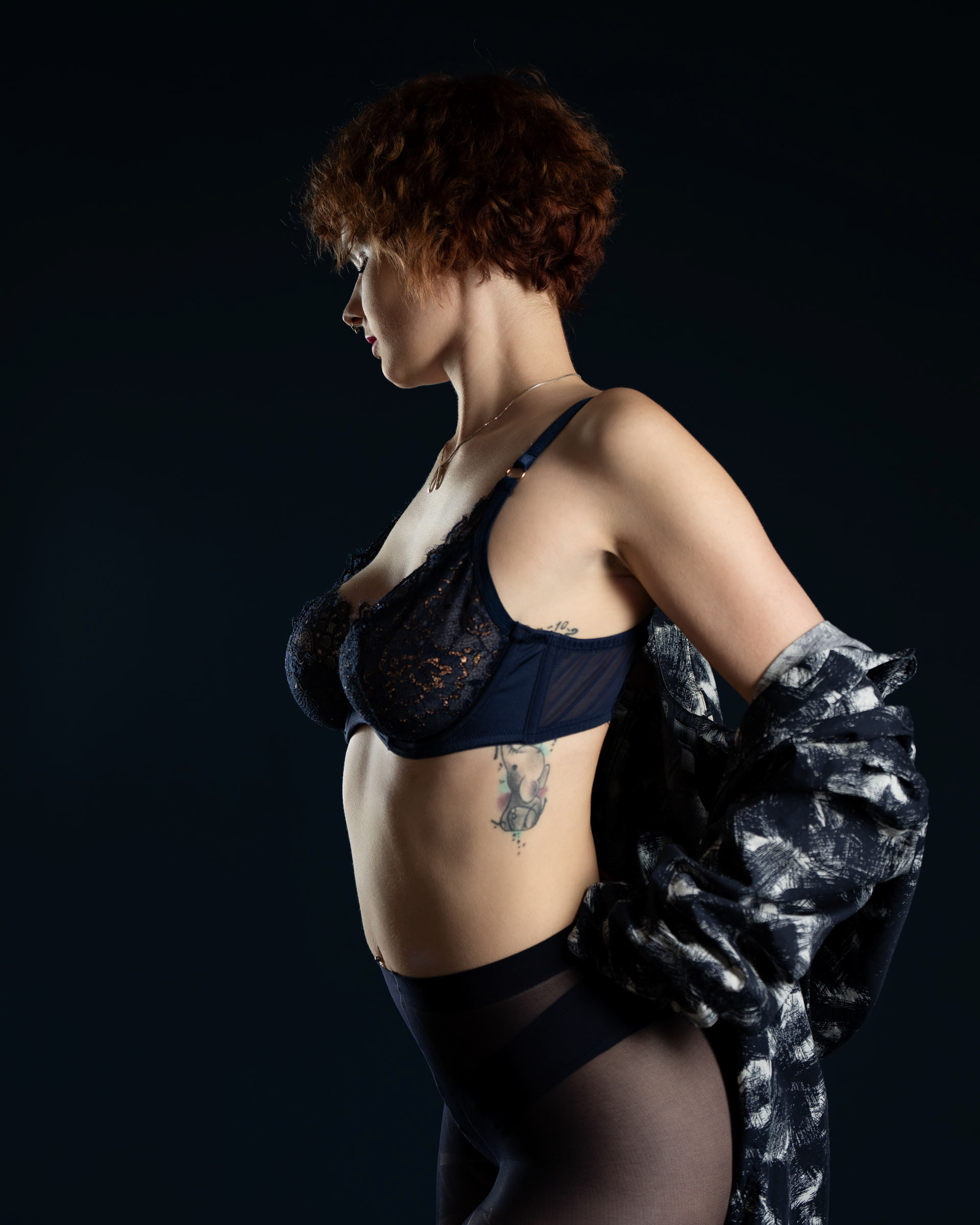 Side profile of Sisse removing shirt, showcasing tattoo and navy lace lingerie in a minimalist dark setting.