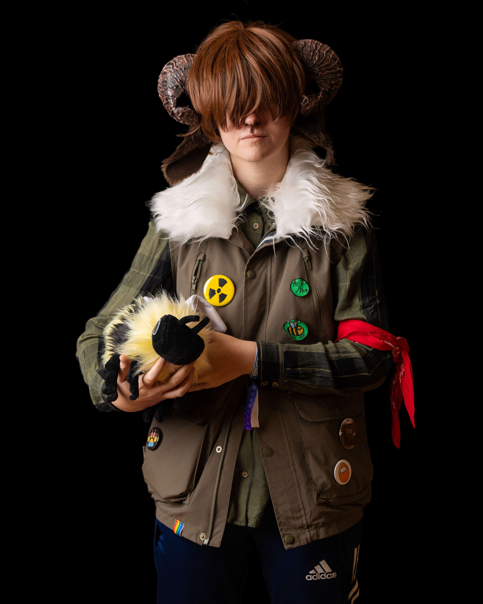 Cosplayer King wearing ram‑style horns and layered costume holding a plush creature, studio portrait with black background.