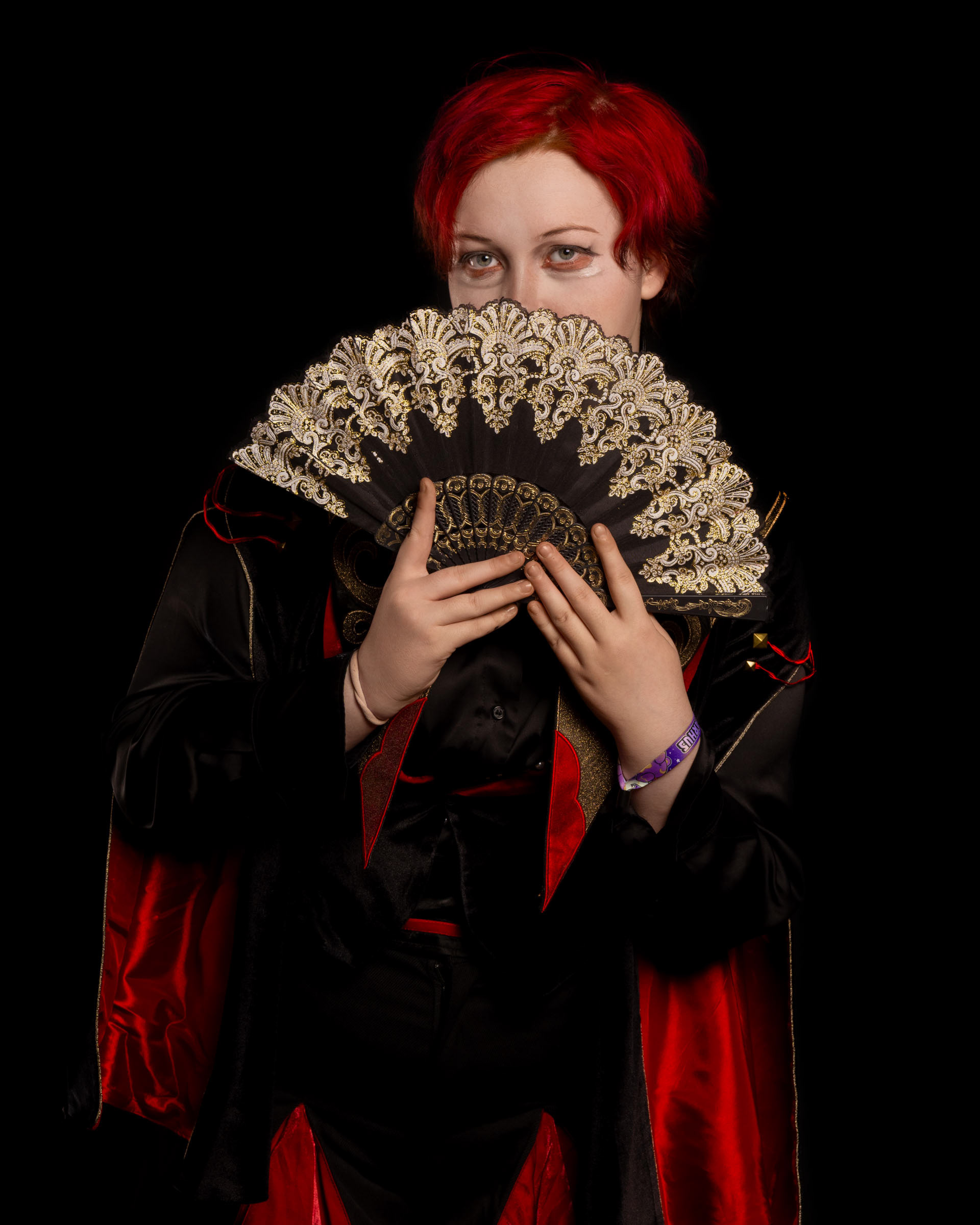 Cosplayer zen.c0z holding a decorative fan up to her eyes, dressed in a black-and-red outfit against a black background.