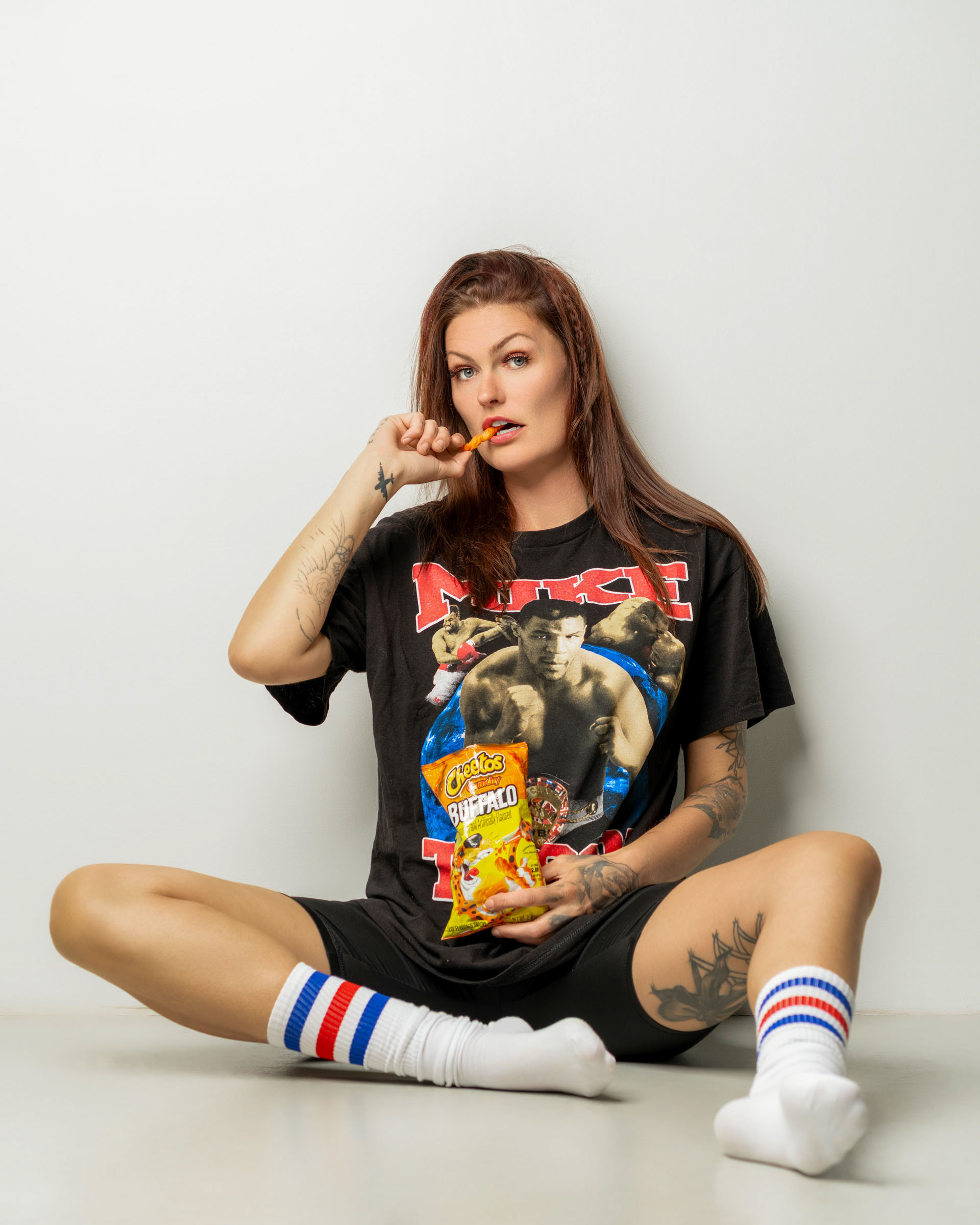 Casual portrait of a woman sitting on the floor eating a Cheetos snack while wearing a relaxed outfit and striped socks.