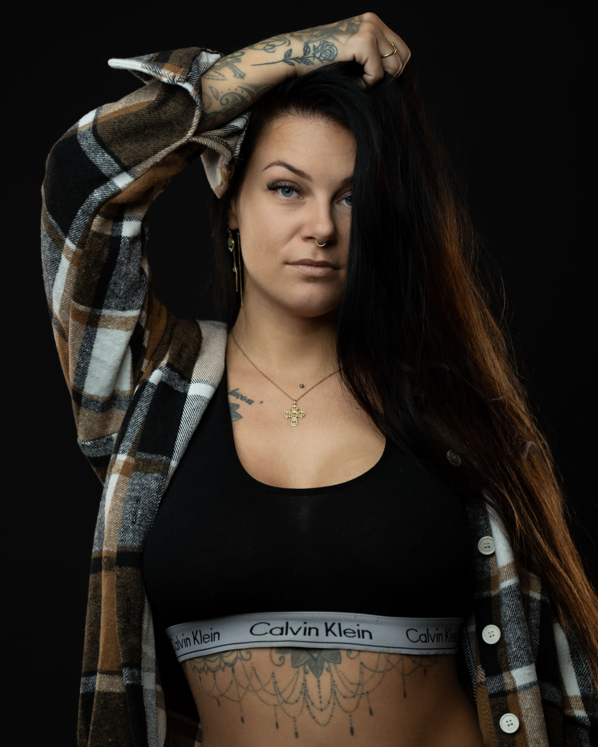 Close-up style studio portrait of Amanda with one arm raised, showing tattoos, plaid shirt and black sports bra.