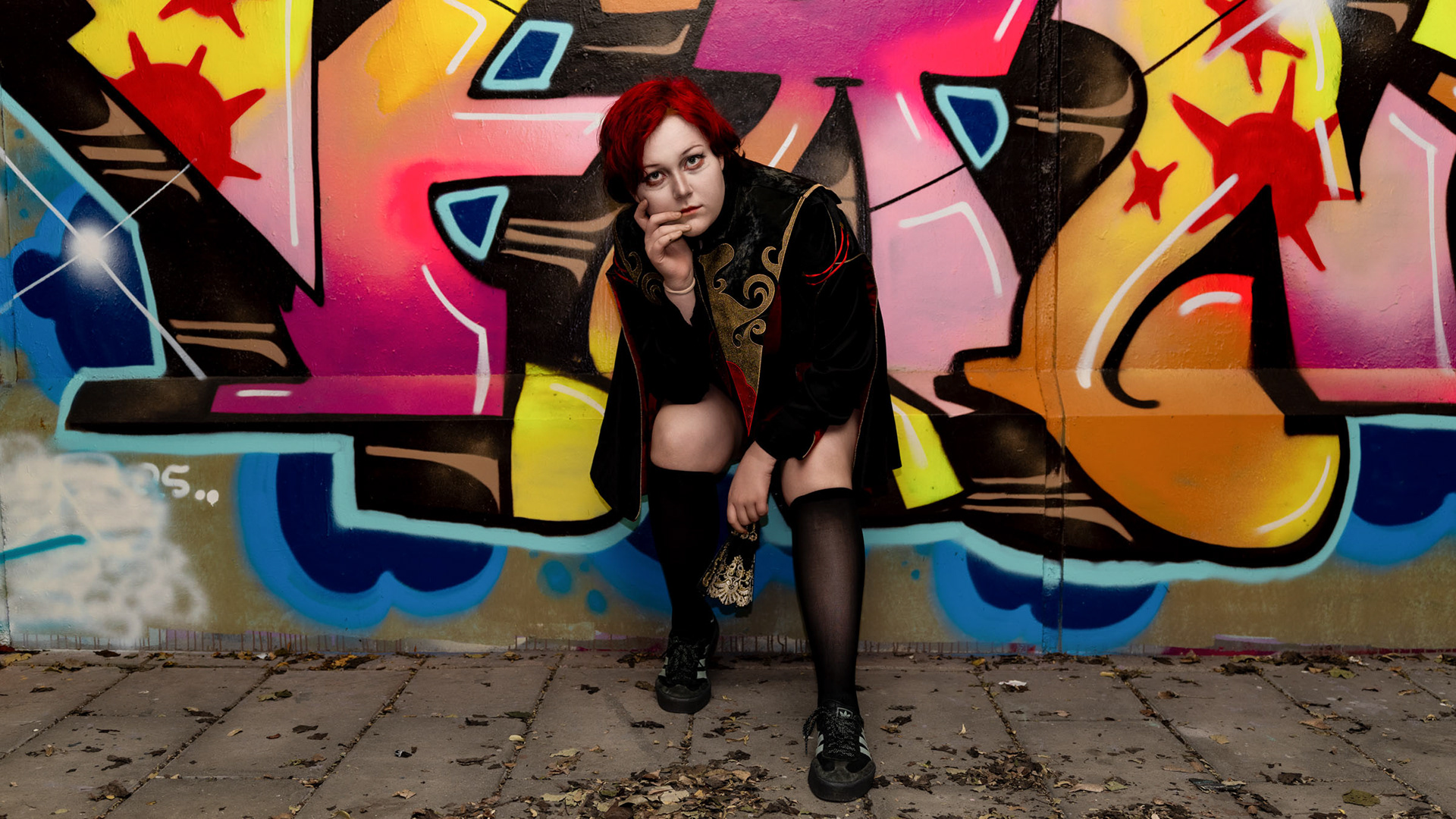 Cosplayer zen.c0z posing in front of a colorful graffiti wall, wearing a black-and-red costume and kneeling with a thoughtful expression.