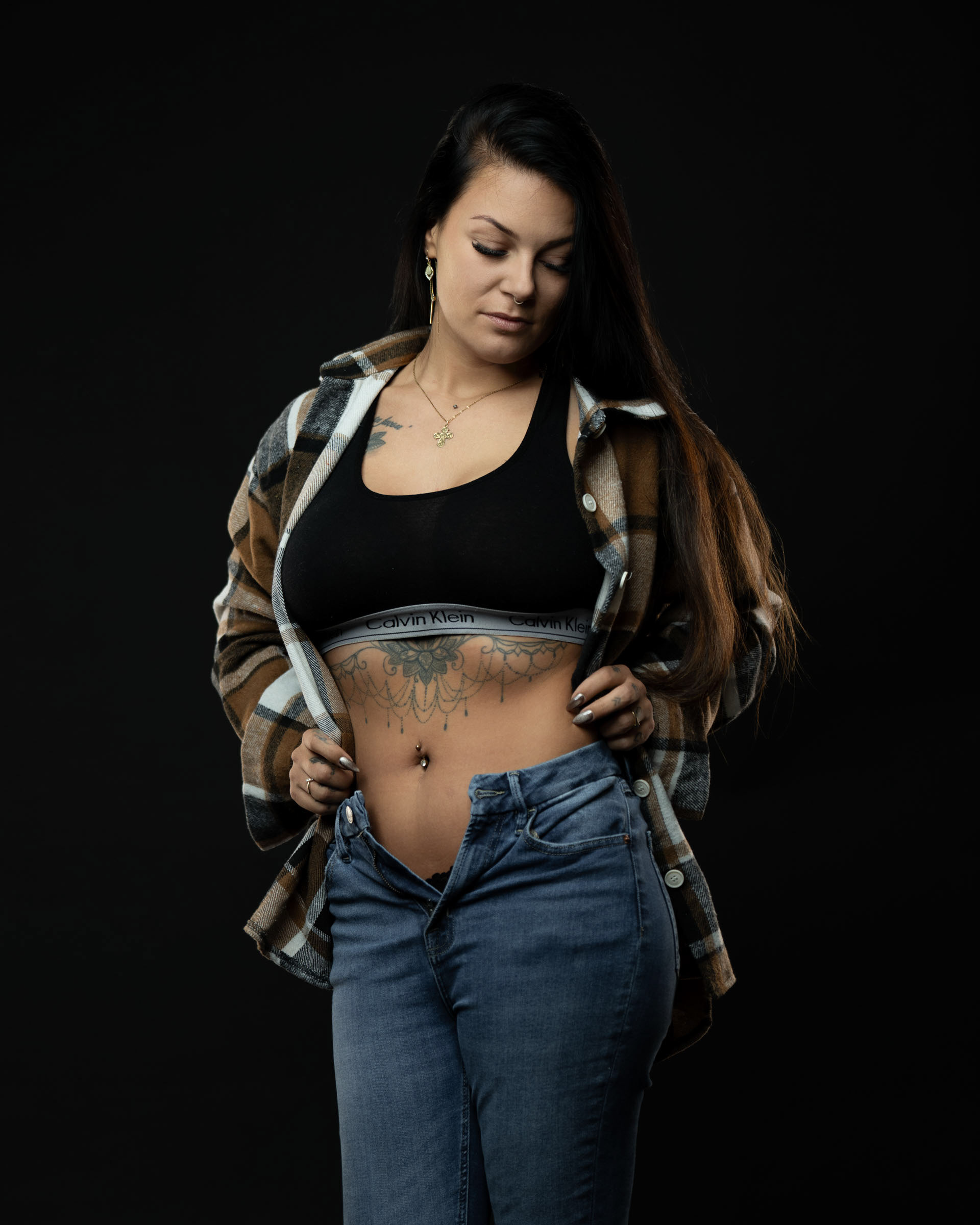 Amanda looking down while slightly holding her shirt open, dressed in a black sports bra and jeans in a dark studio setup.