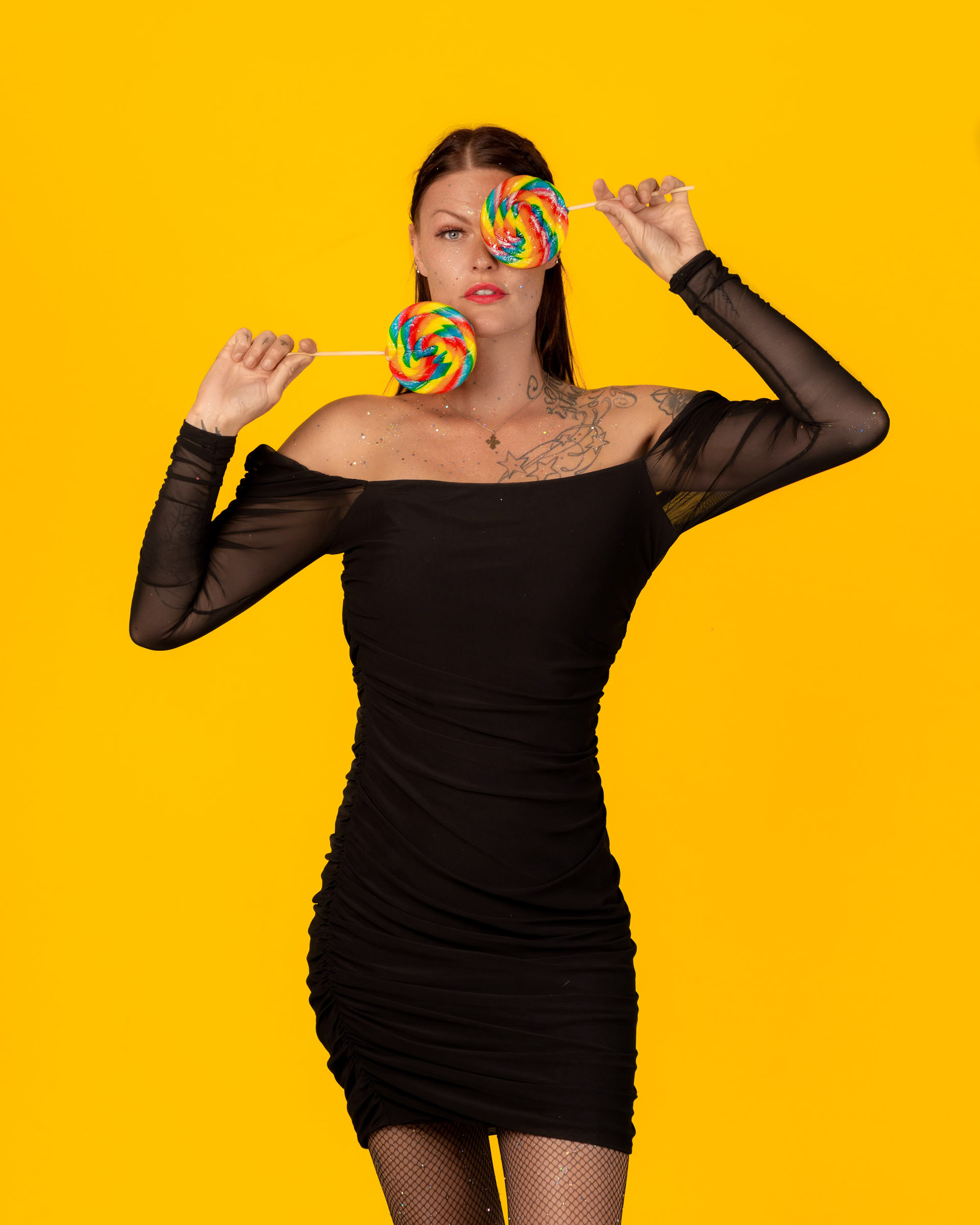 Creative candy-themed pose with a model covering one eye using a rainbow lollipop while standing confidently in a black dress.