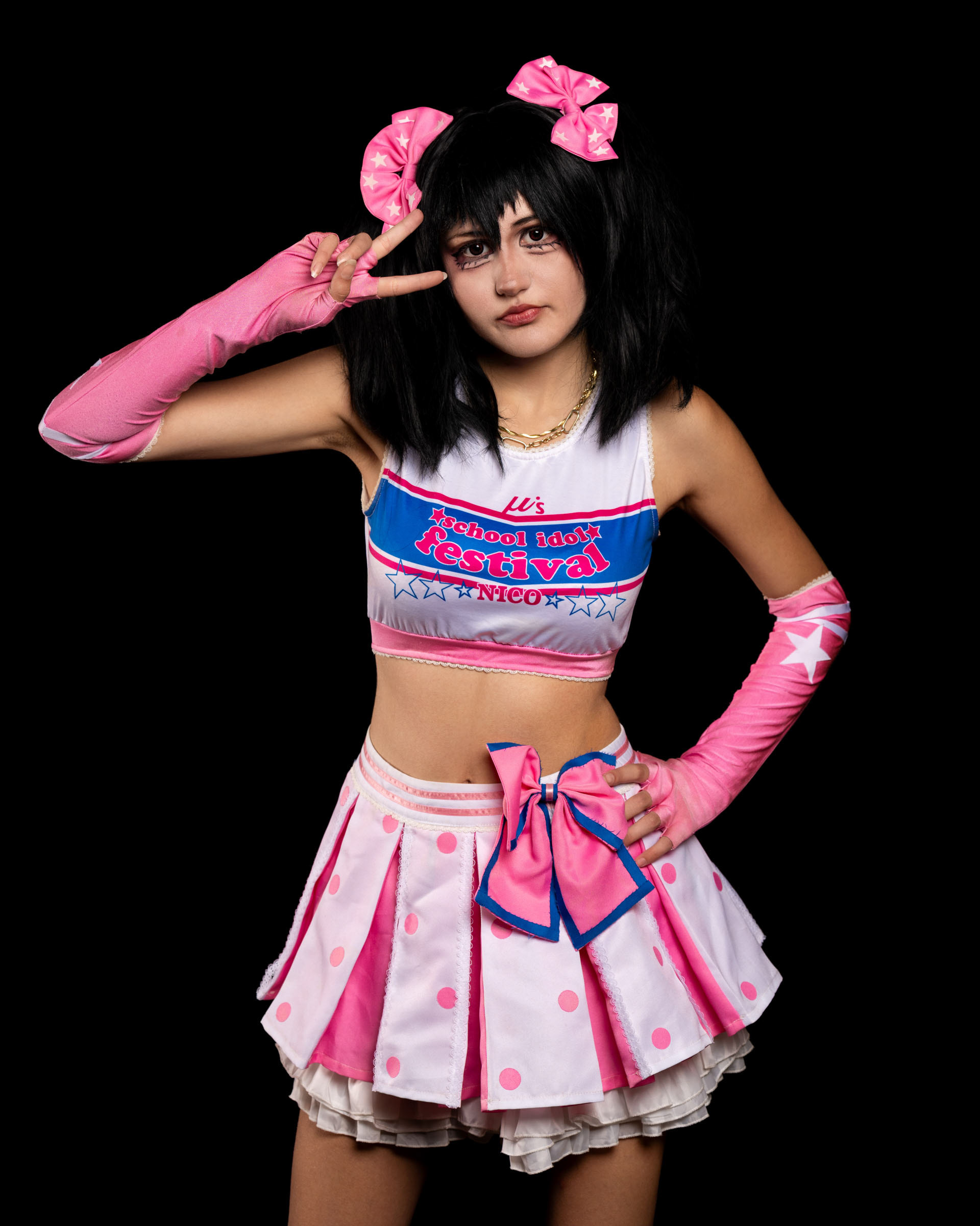 Studio cosplay shot of may_c0z in Nico Yazawa attire, posing with a hand on her head and expressing character attitude.