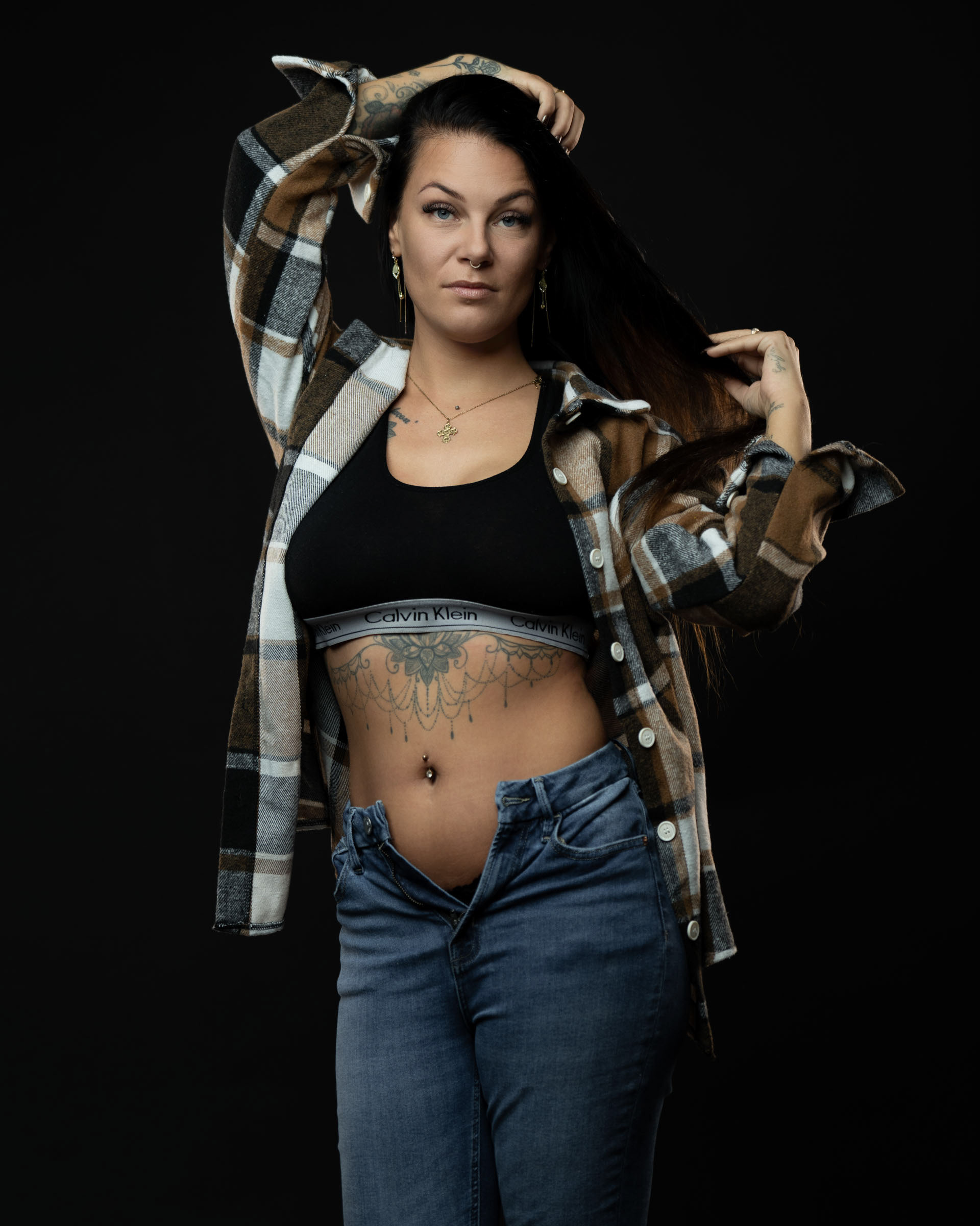 Studio portrait of Amanda lifting her hair with one hand, wearing a plaid shirt, black sports bra and open jeans