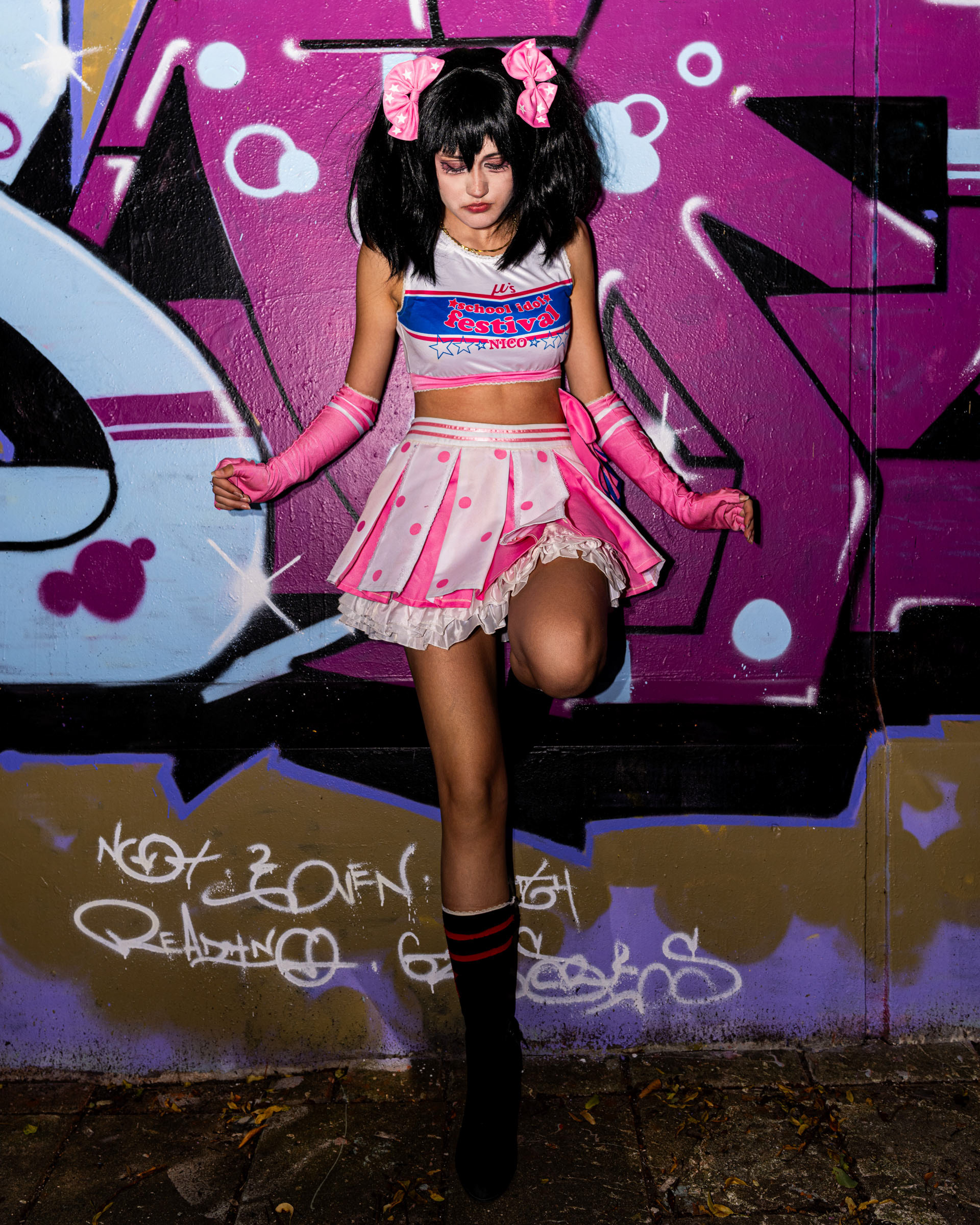Full-body outdoor photo of may_c0z as Nico Yazawa, posing with one knee raised in front of a bright graffiti background at FanCon Aarhus.
