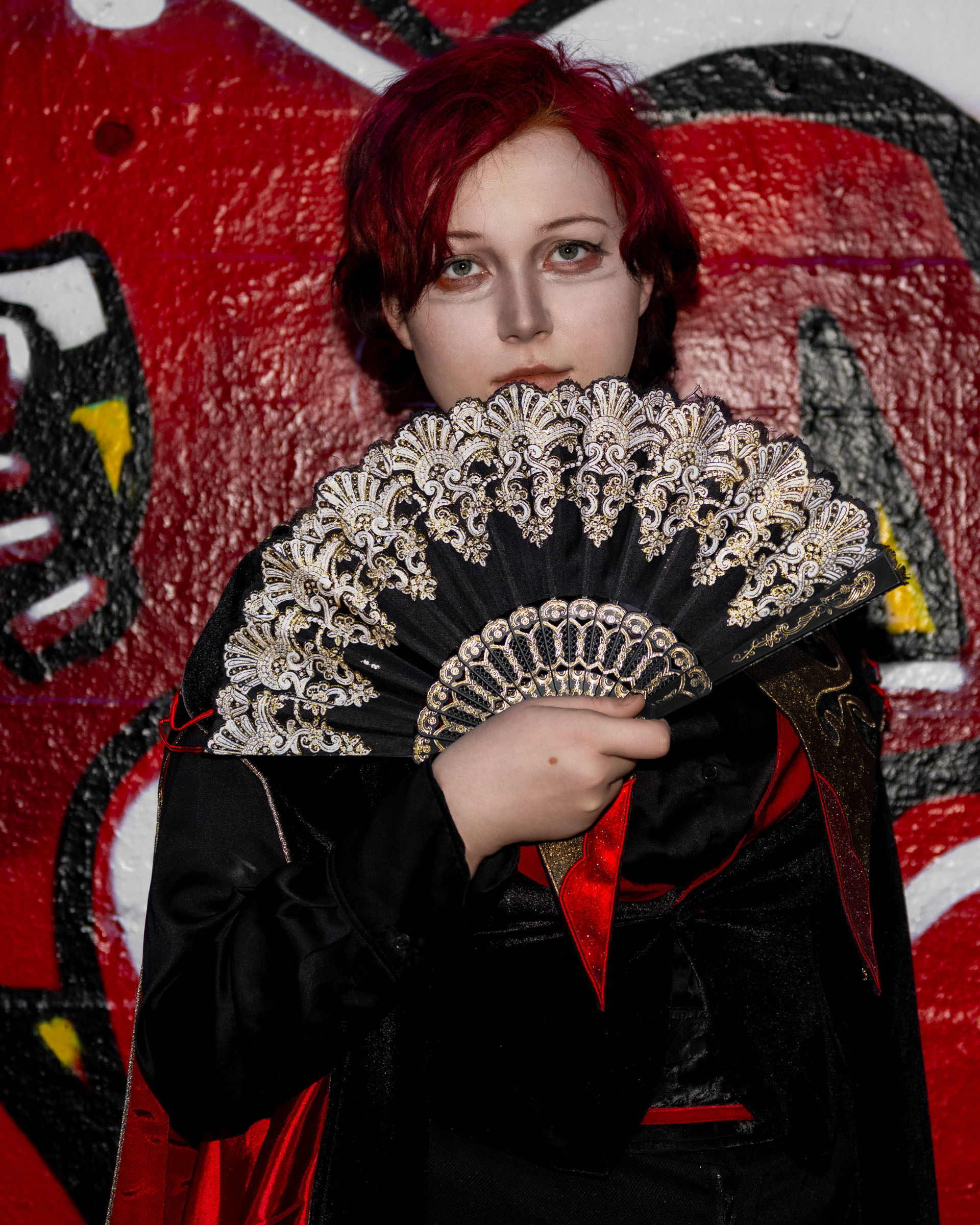 Close-up of cosplayer zen.c0z holding an ornate folding fan partly covering her face, with red graffiti artwork behind her.