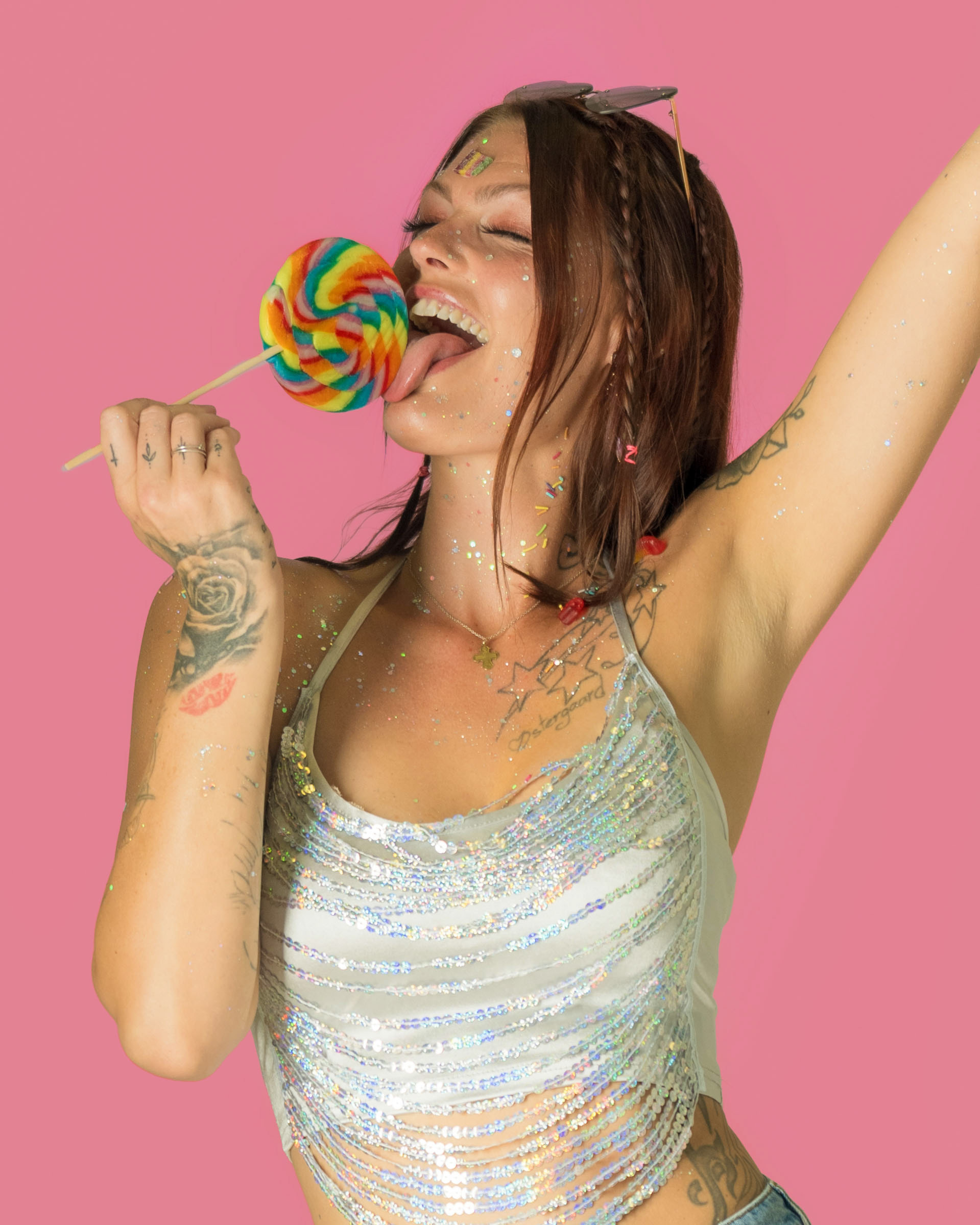 Female model licking a colorful rainbow lollipop against a bright pink background, covered in glitter for a playful candy-themed look.