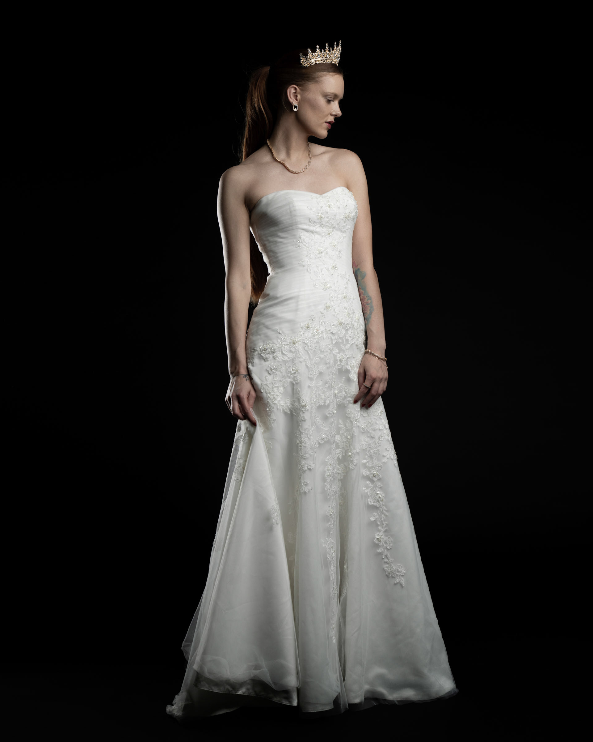Side view of Noamynte in a strapless wedding dress and crown, looking down in a graceful posture.