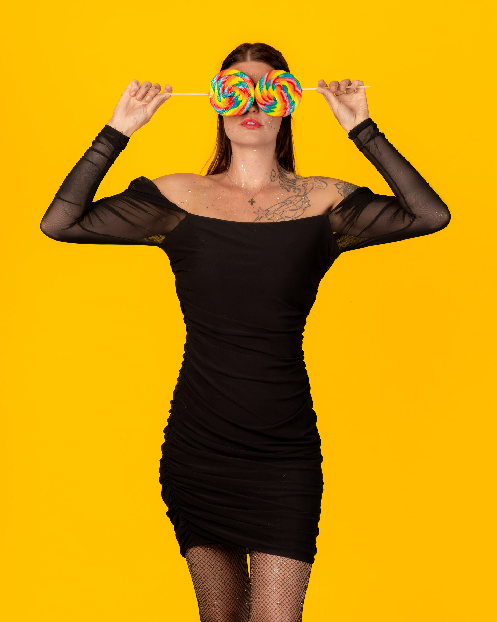Model in a black dress holding two rainbow lollipops over her eyes in a colorful, candy-inspired portrait on a yellow background.