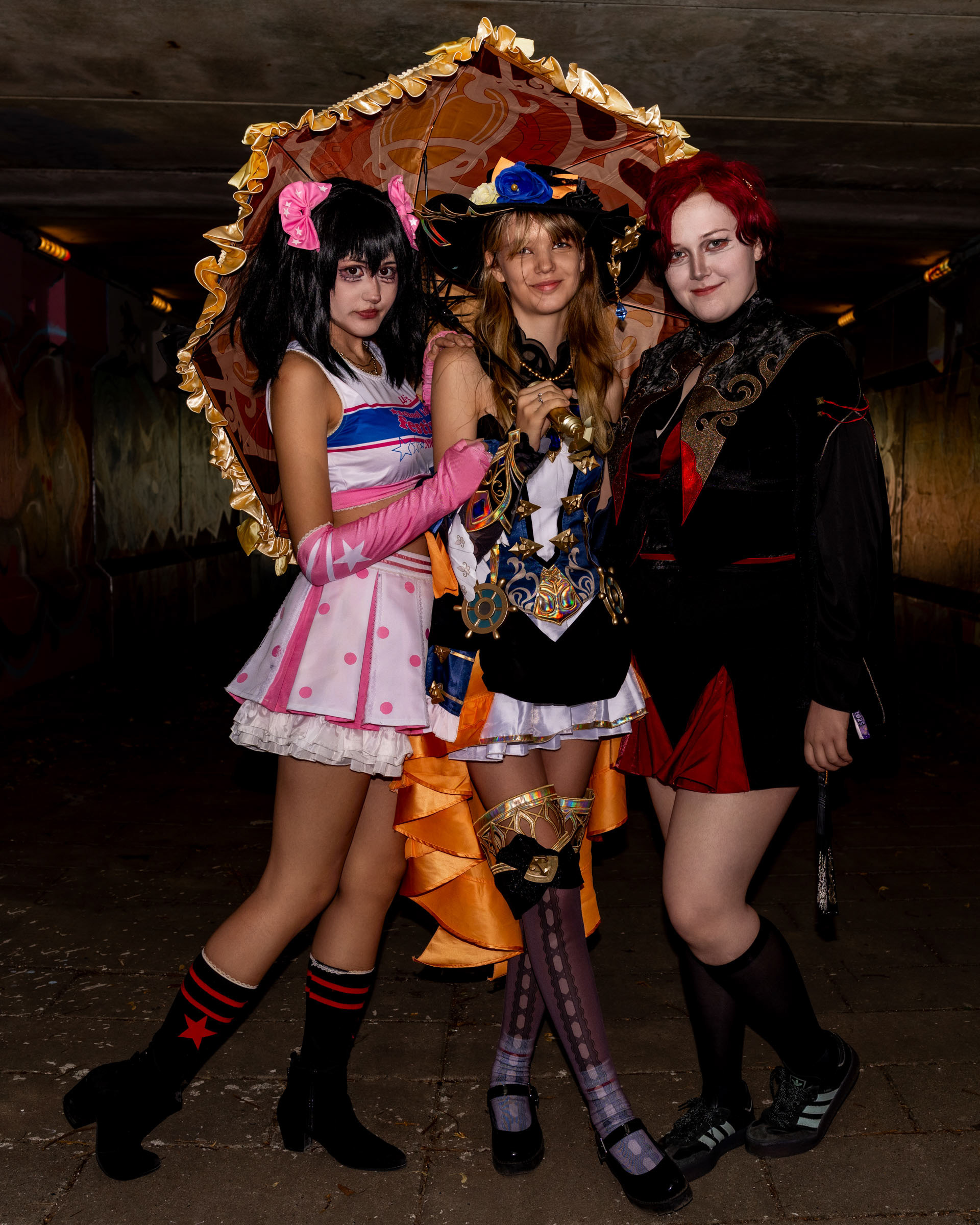 Cosplayers may_c0z, ruxqym and zen.c0z standing together under a decorative parasol, dressed in colorful costumes and posing in an outdoor tunnel.