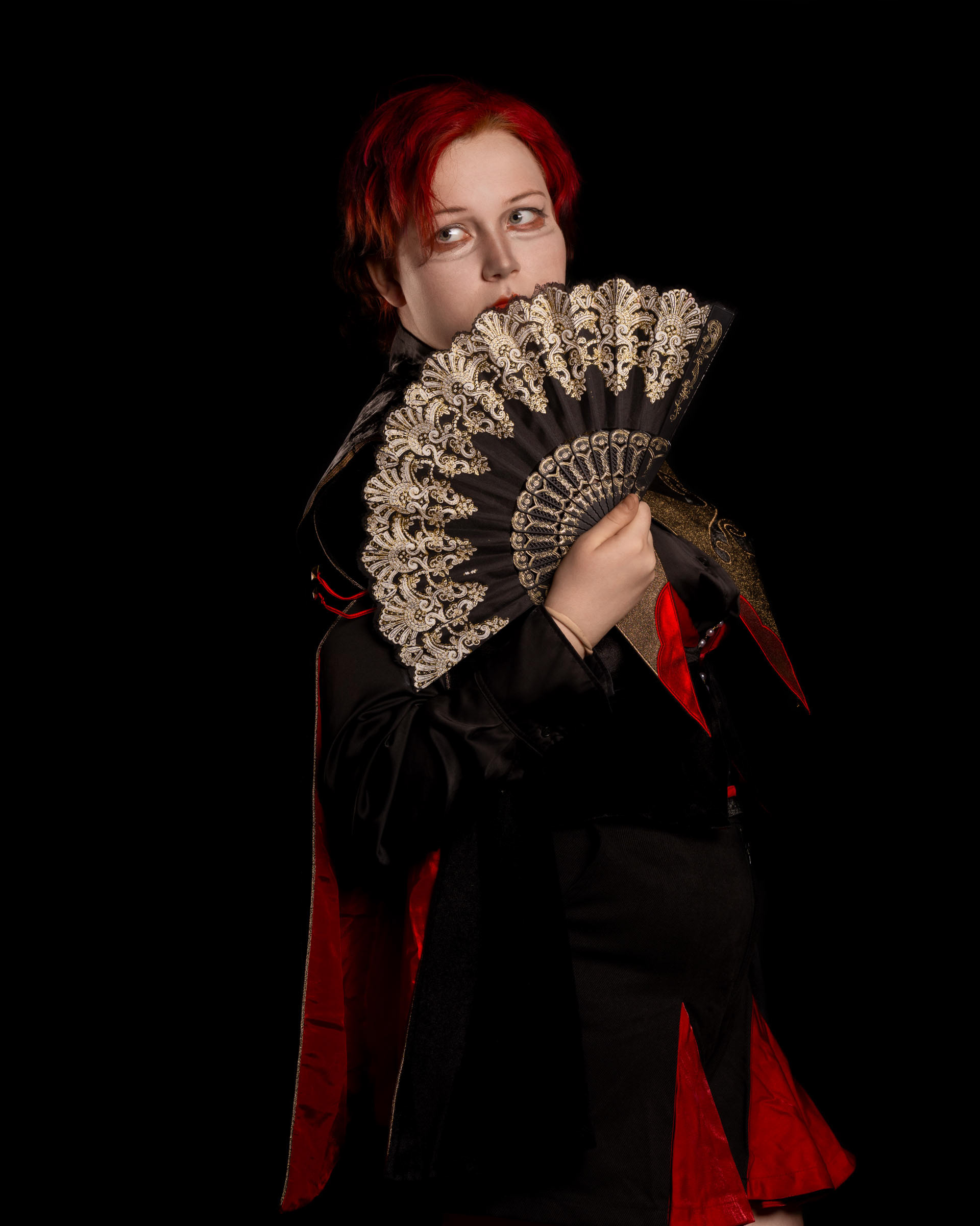 Cosplayer zen.c0z posing with an ornate fan while looking to the side, dressed in a black and red costume against a dark backdrop.