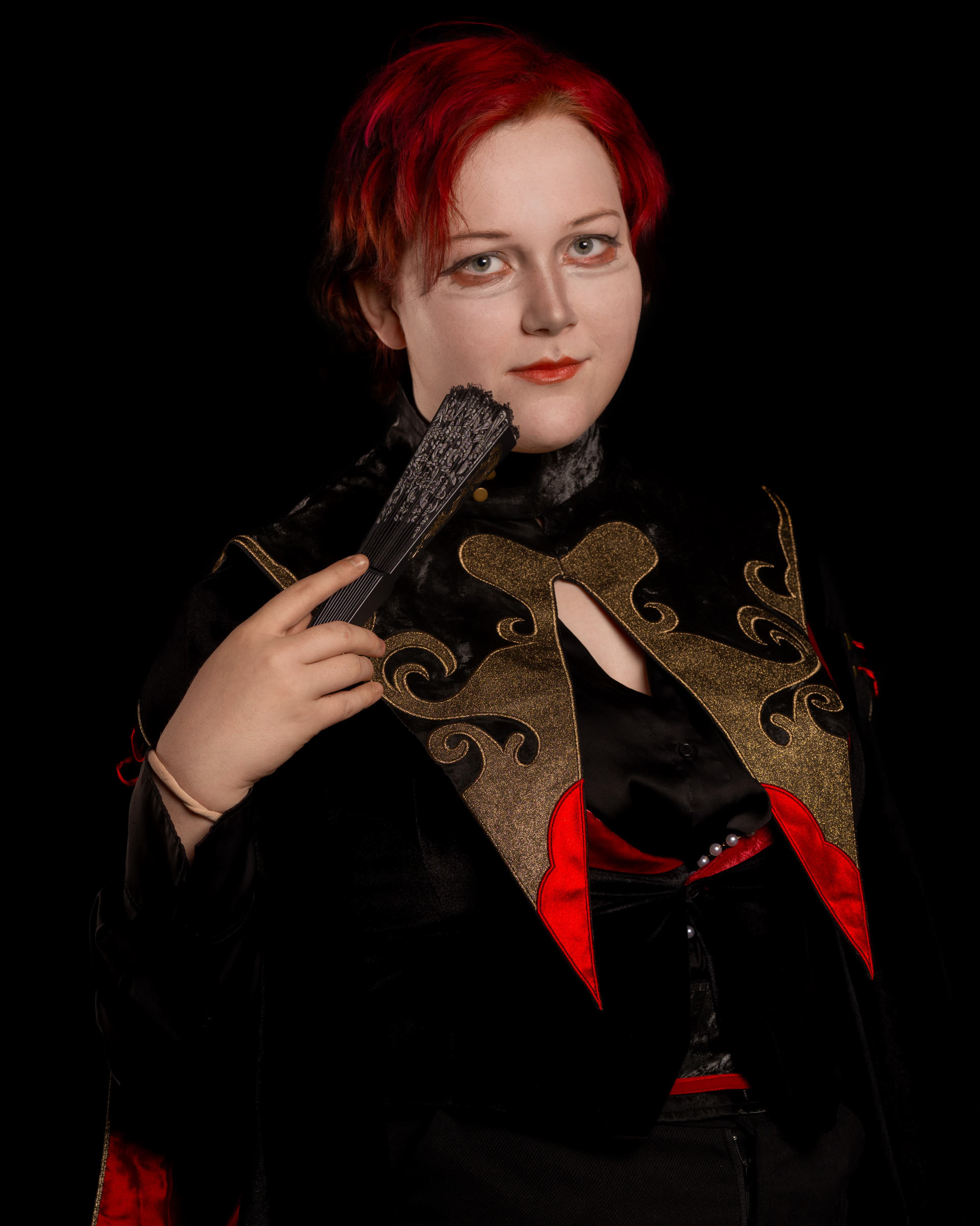 Portrait of cosplayer zen.c0z holding a textured folding fan and wearing a detailed black-and-red costume against a black background.