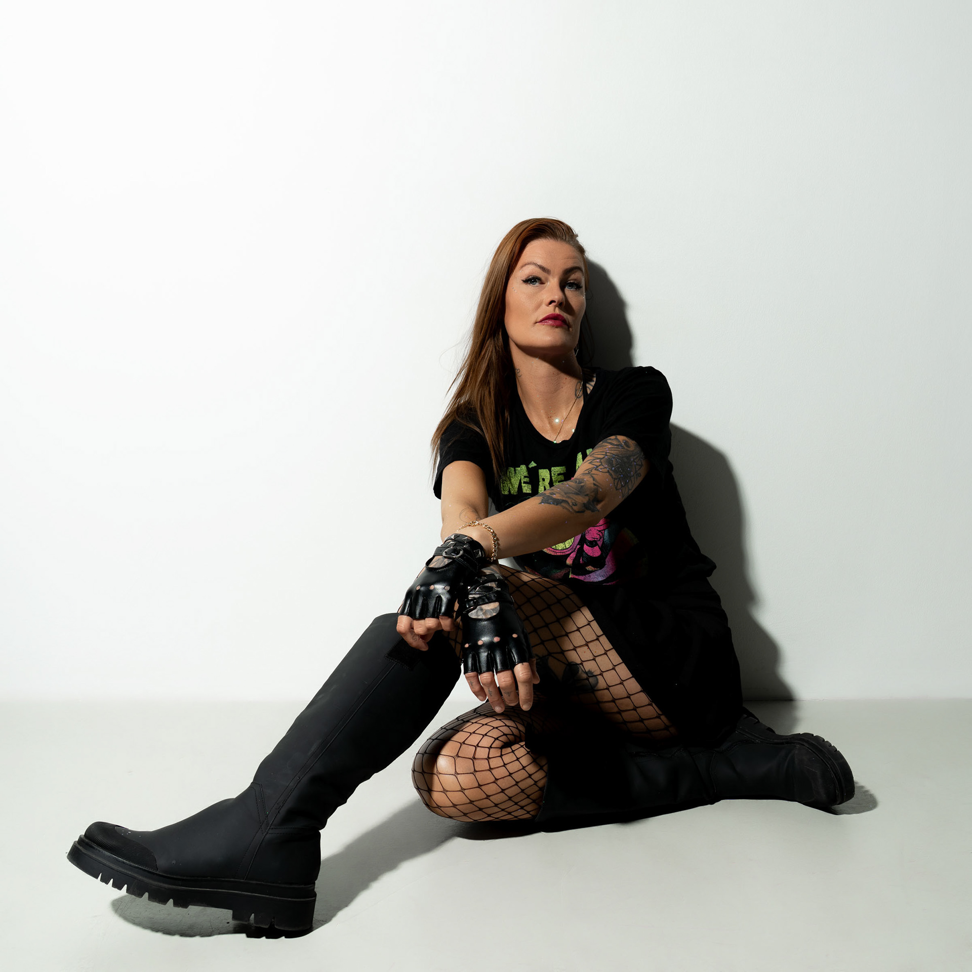 Simone seated on the floor with legs stretched, leaning slightly forward, her gloves and tattoos visible.