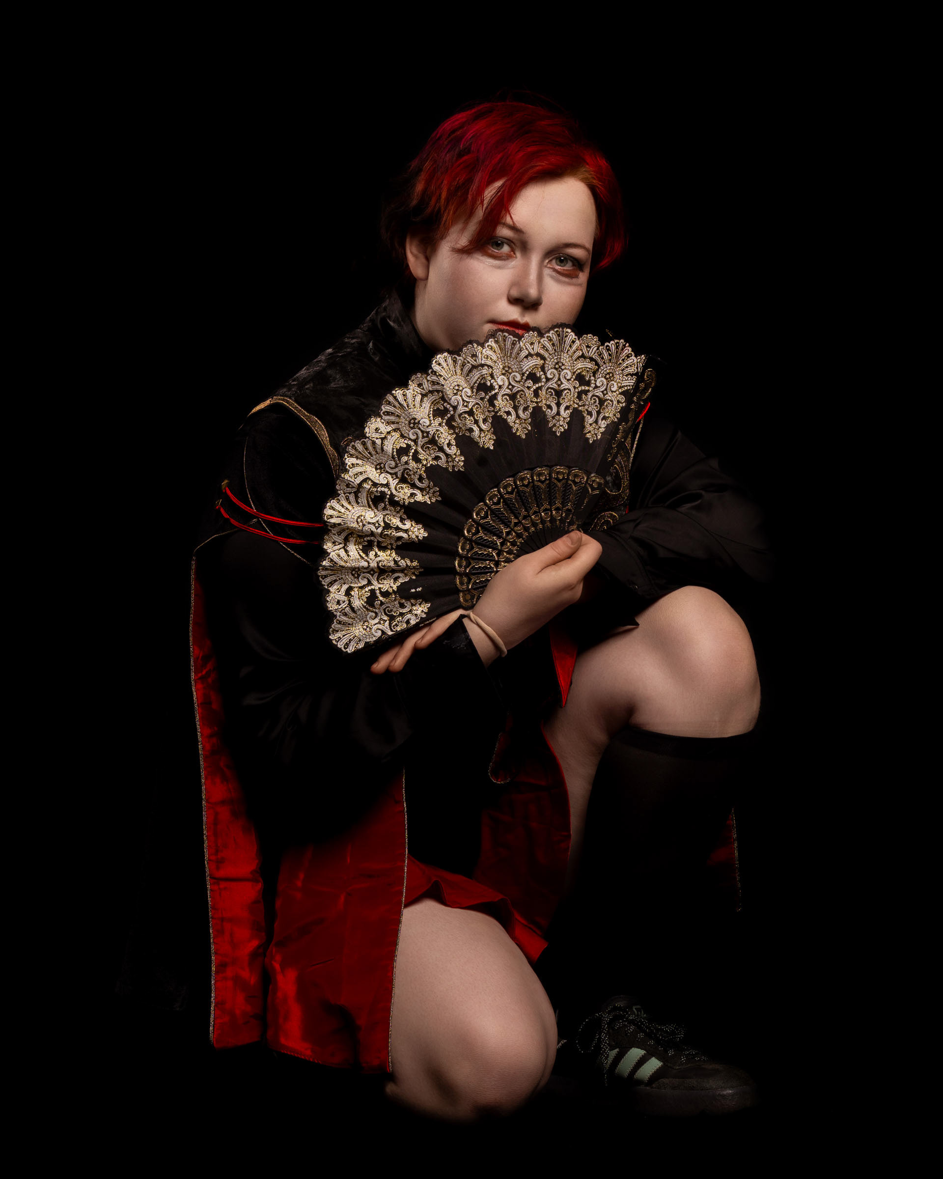 Cosplayer zen.c0z kneeling while holding a black-and-gold decorative fan, wearing a cape with red lining against a dark background.