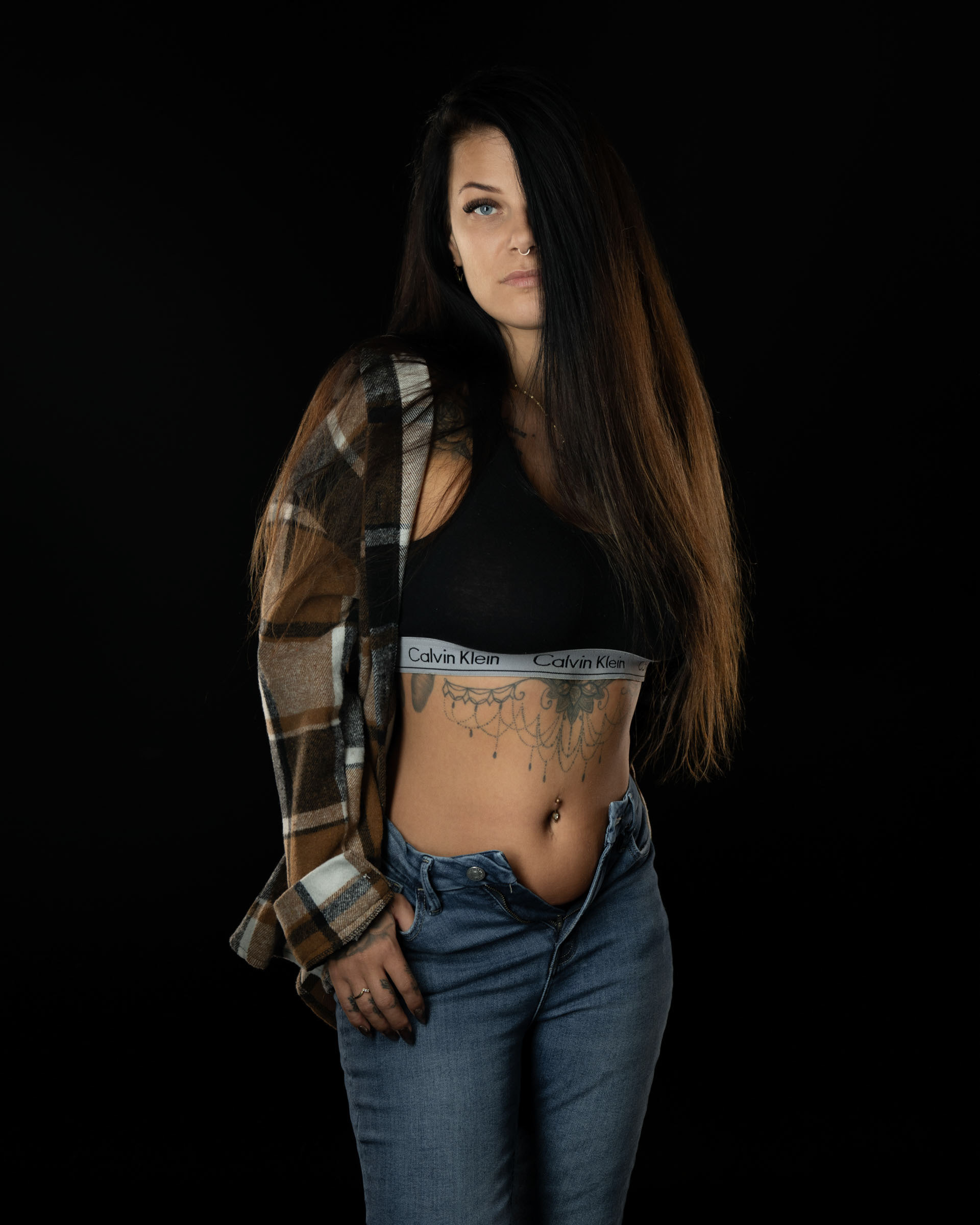 Amanda posing with long hair covering part of her face, wearing a plaid shirt, black sports bra and jeans in a dark studio environment.