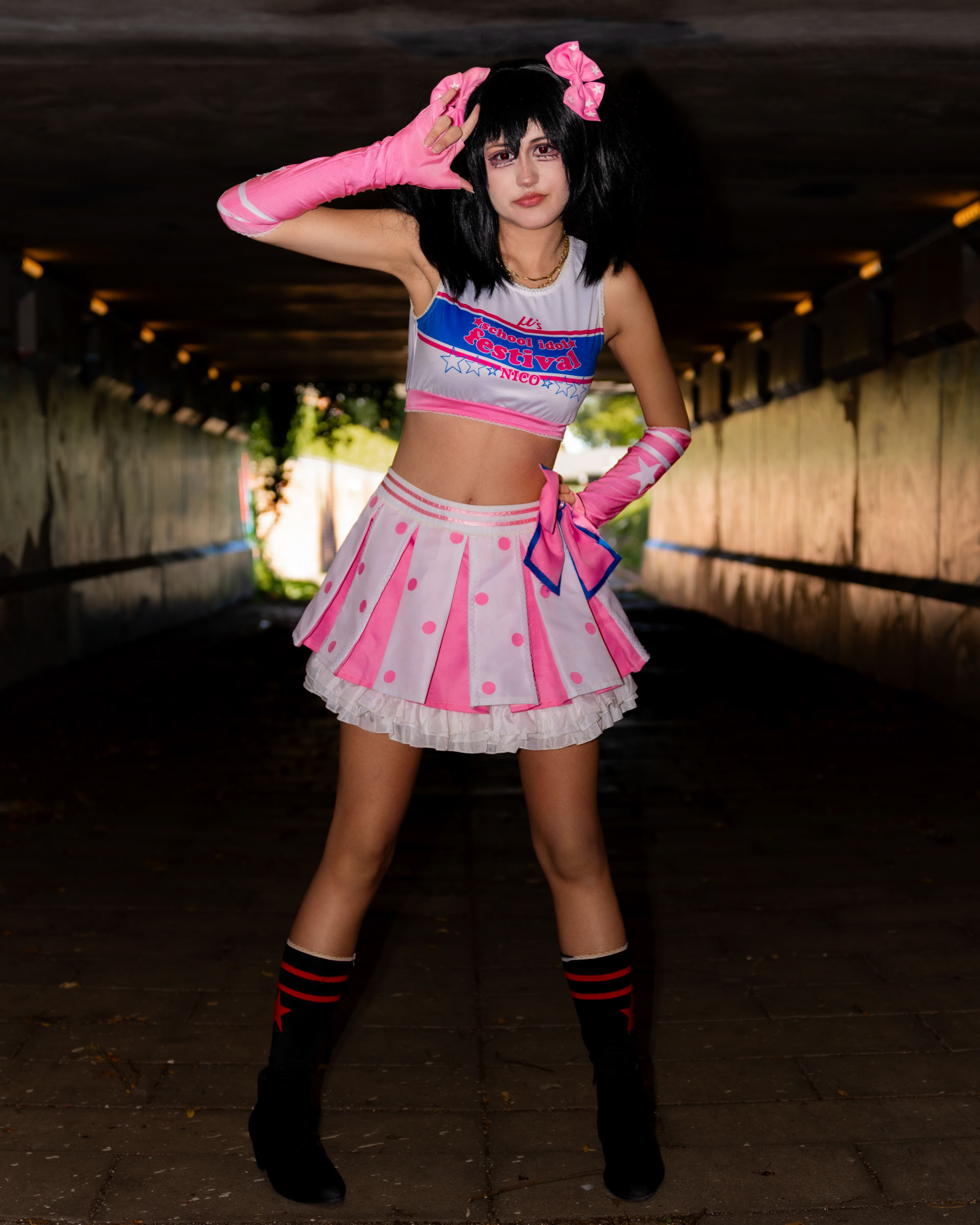 Full-body photo of cosplayer may_c0z dressed as Nico Yazawa, posing with a peace sign in a tunnel, wearing a pink-and-white idol costume with dramatic flash lighting and dark surroundings.