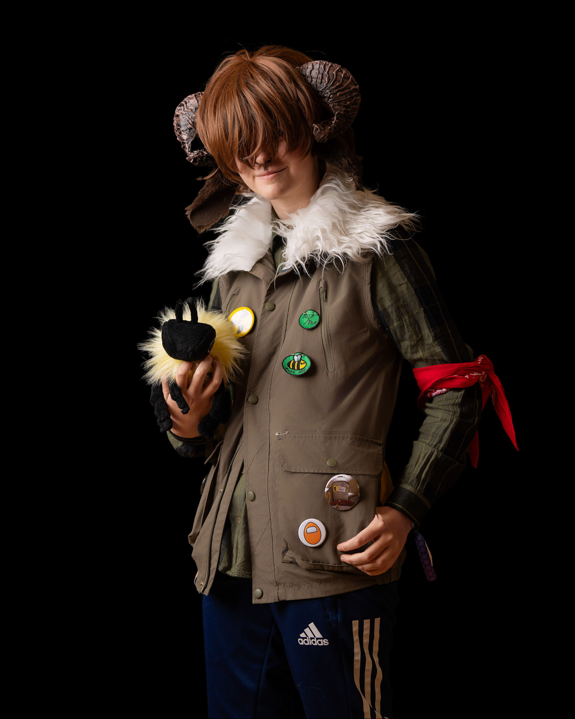 Creative cosplay portrait of King with horn headpiece and textured vest covered in badges, holding a fuzzy plush toy in both hands.