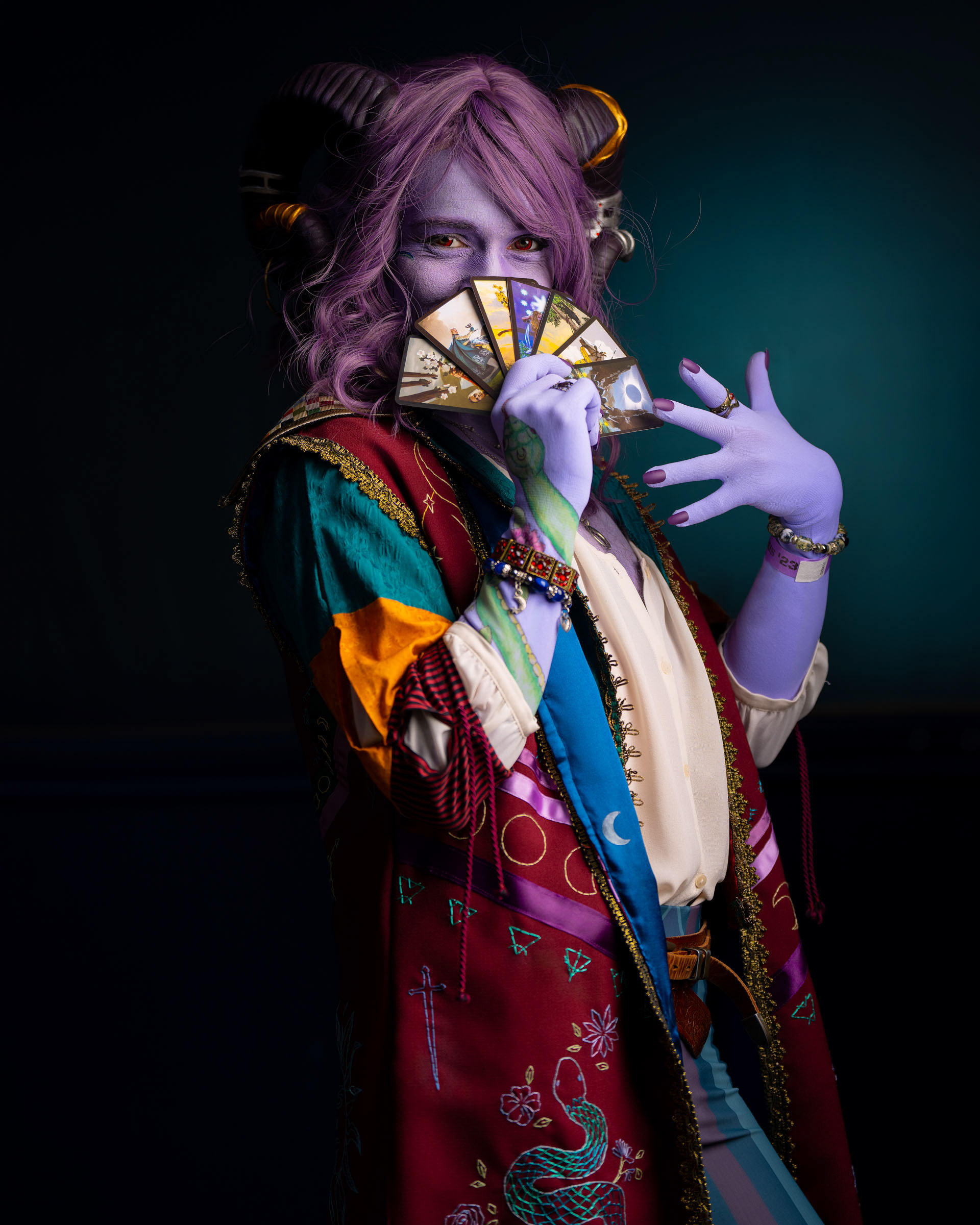 Jester Lavorre cosplay holding tarot cards in front of face, purple skin and multicolored outfit, playful expression and dramatic lighting.
