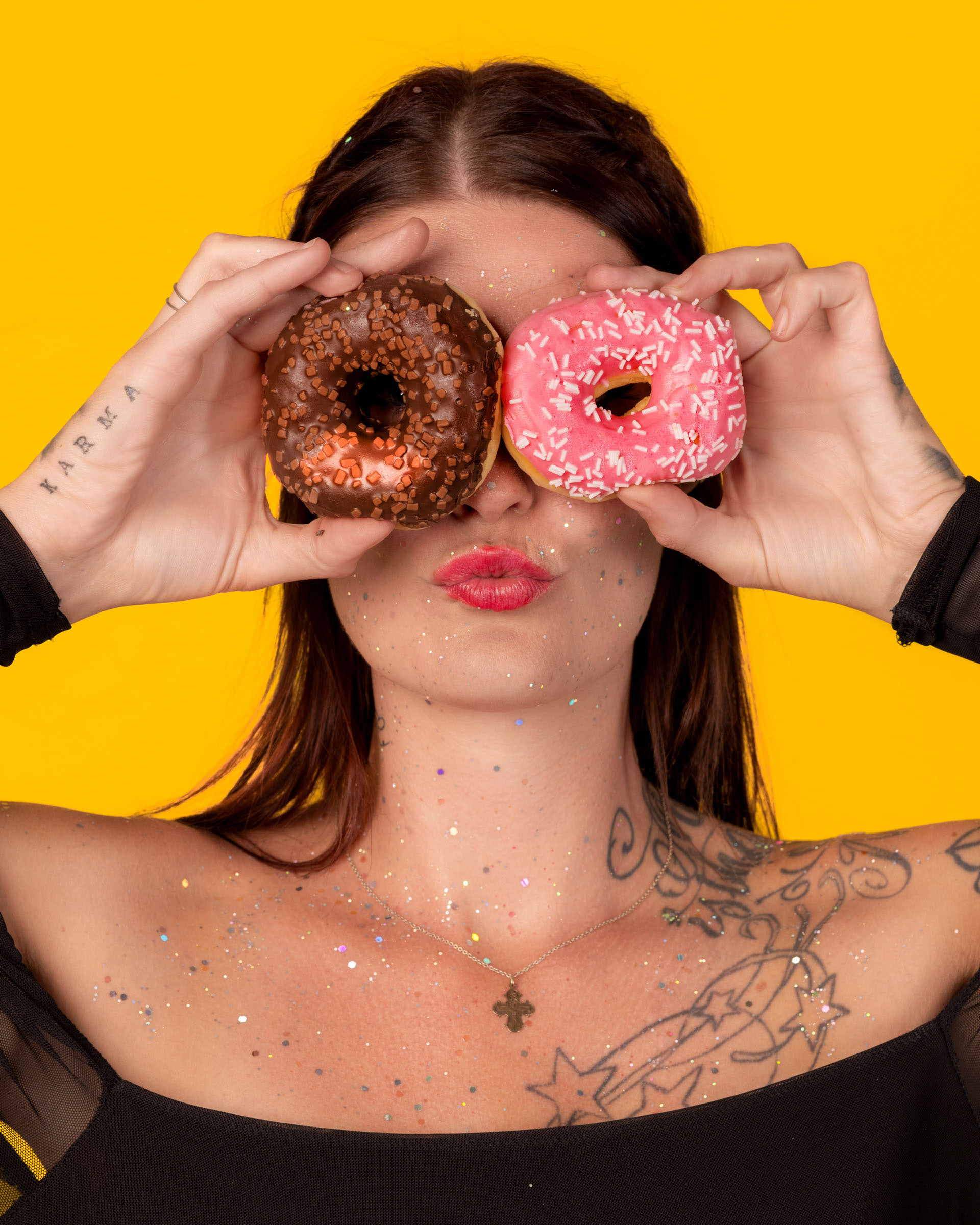 Playful portrait of a woman holding two donuts, one in front of her eye, against a bright yellow studio background.
