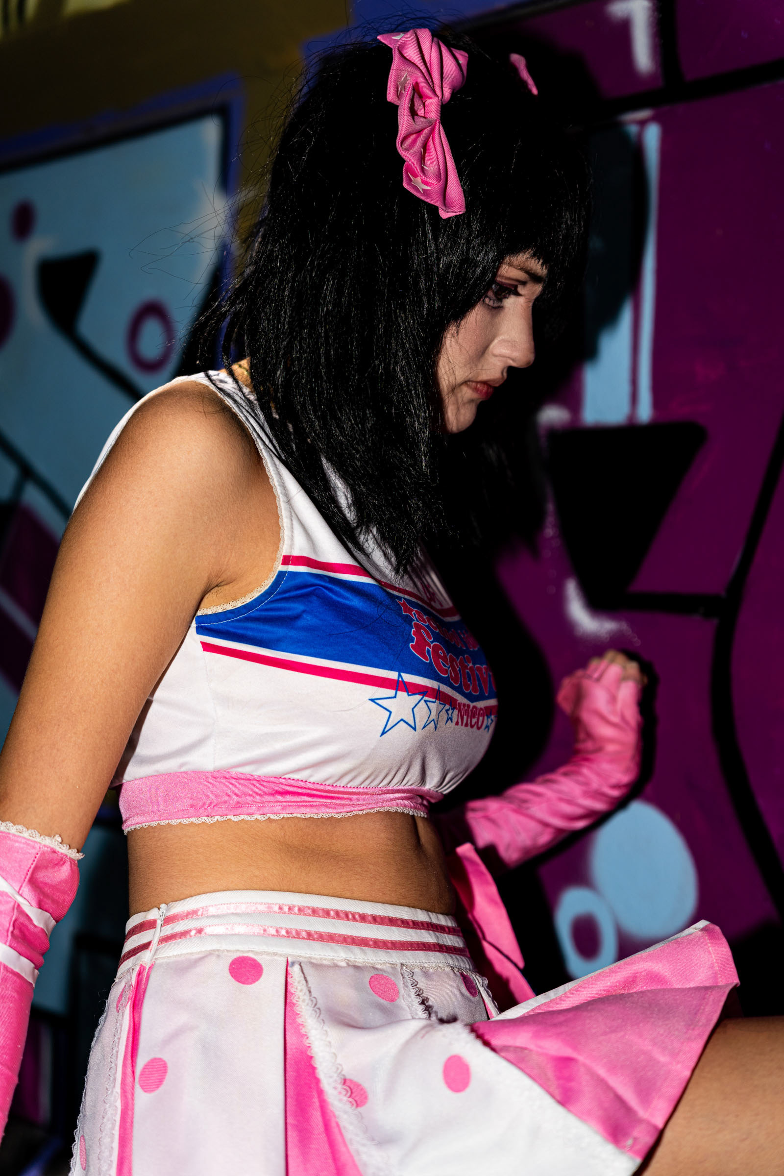Close-up side view of cosplayer may_c0z dressed as Nico Yazawa, standing against a purple graffiti wall, with focus on the pink-and-white idol costume and dramatic flash lighting.