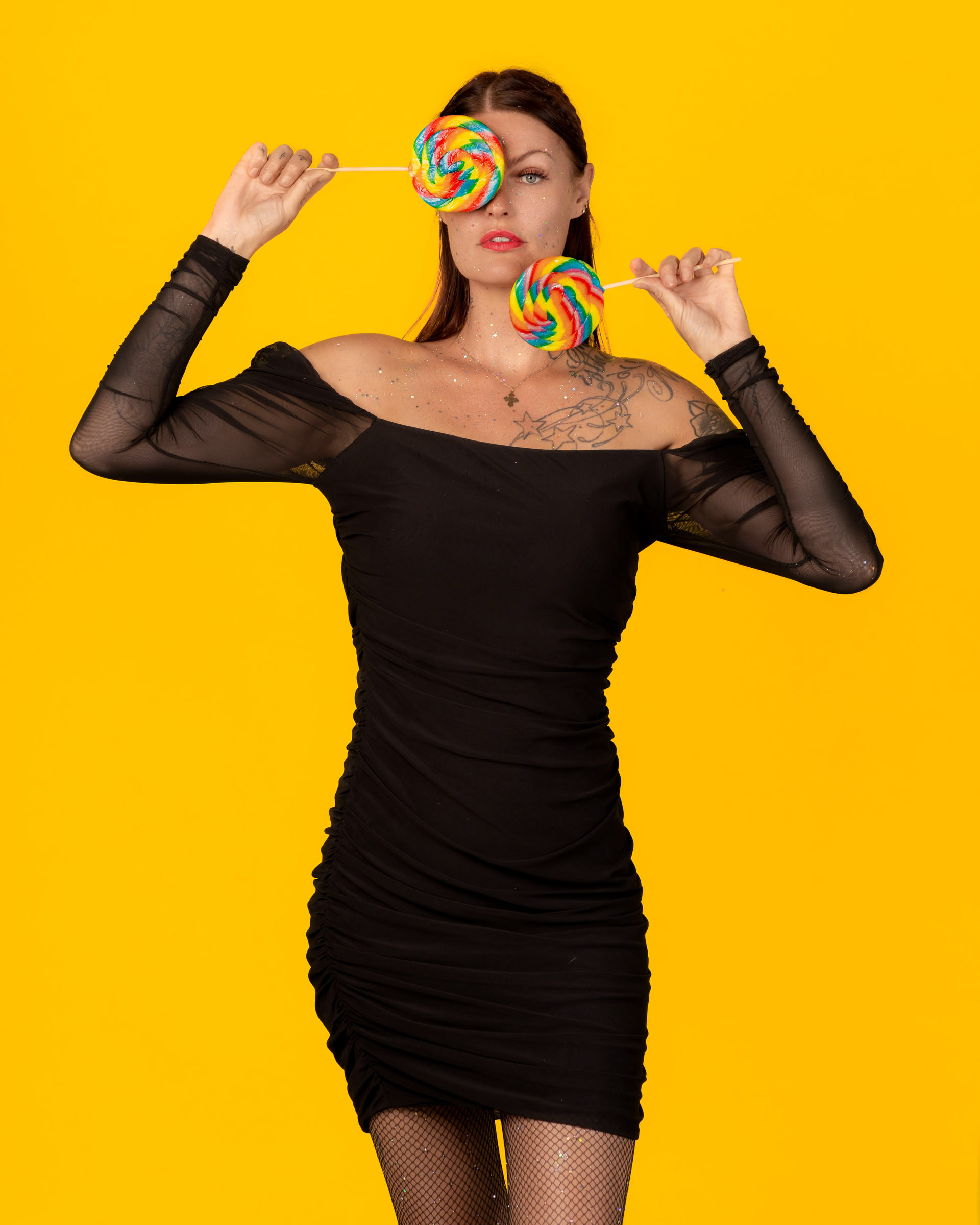 Playful fashion portrait of a woman posing with bright lollipops against a solid yellow backdrop.