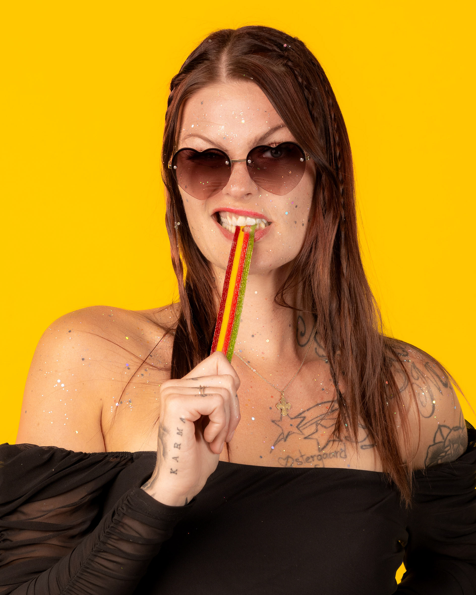 Candy-themed studio portrait featuring a woman posing with sour candy and heart-shaped sunglasses in front of a bright yellow backdrop.