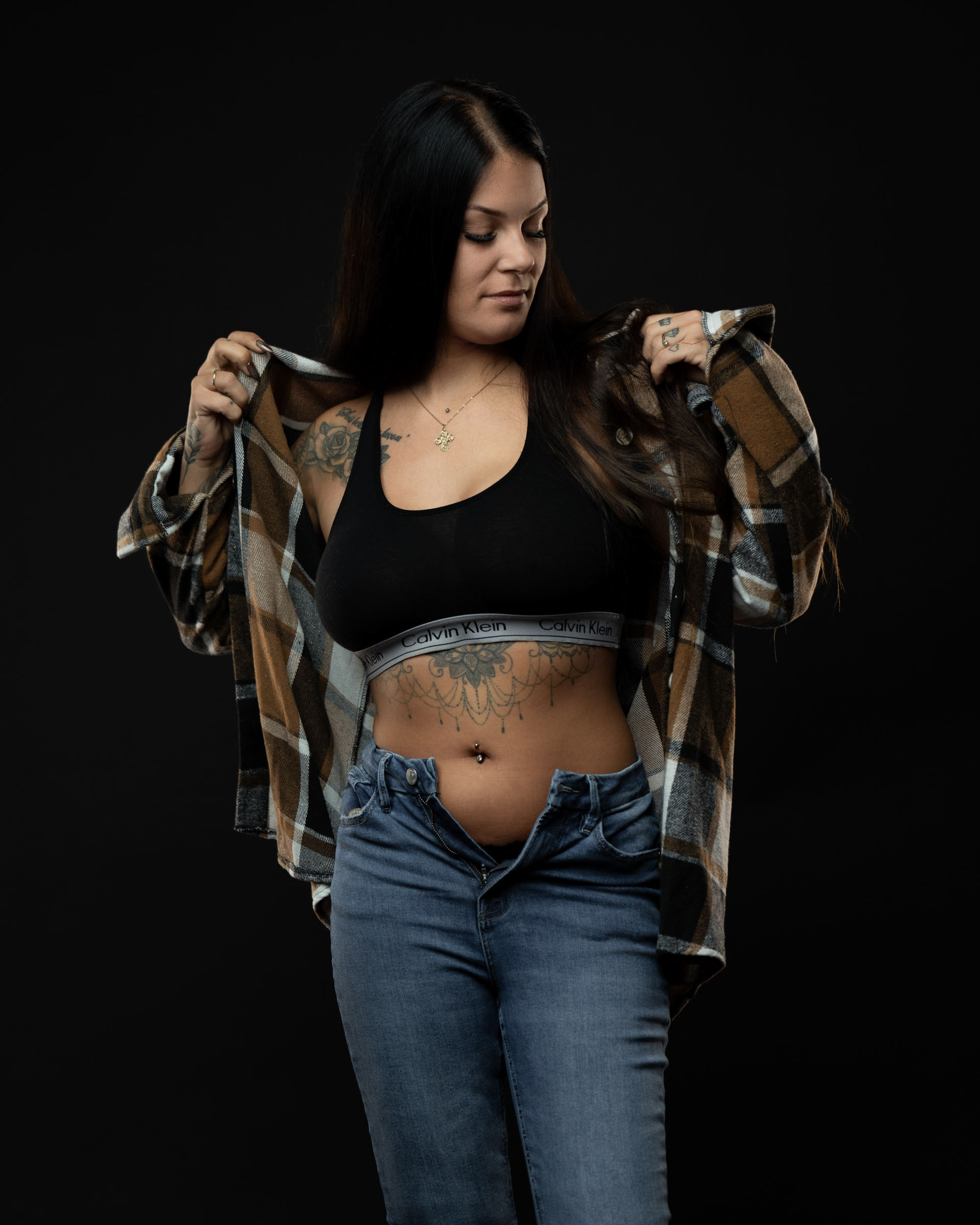 Amanda adjusting her plaid shirt while wearing a black sports bra and open jeans, captured in soft studio lighting.