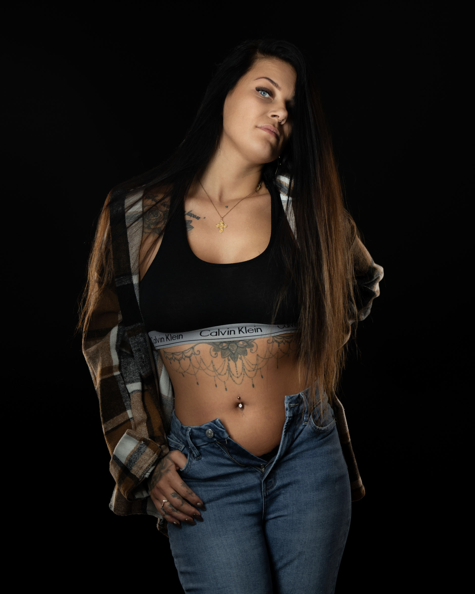 Amanda tilting her head slightly, posing in a plaid shirt and black sports bra with open jeans in a soft-lit studio portrait.
