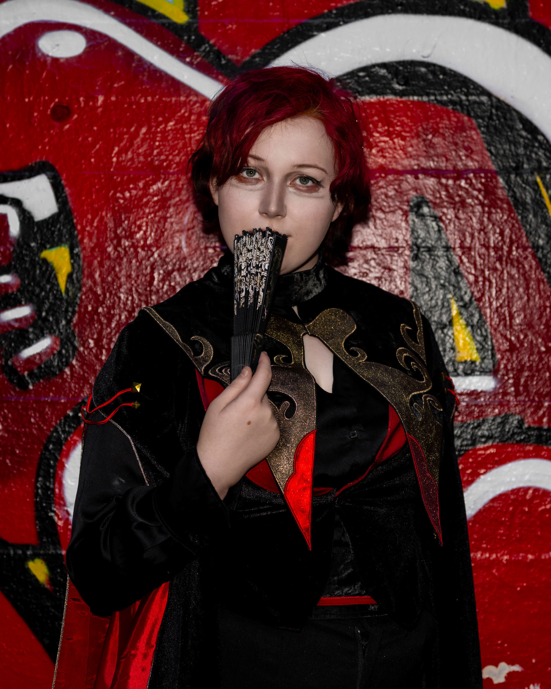 Close-up of cosplayer zen.c0z holding an ornate folding fan partly covering her face, with red graffiti artwork behind her.