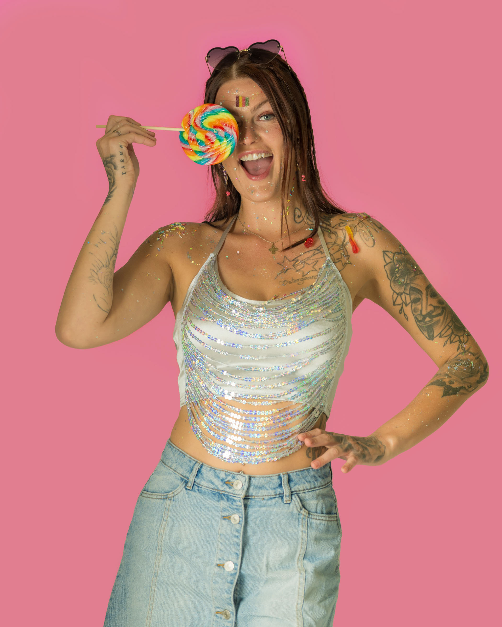 Glitter-covered model posing joyfully with a rainbow lollipop in a colorful, sweet-inspired studio setup.