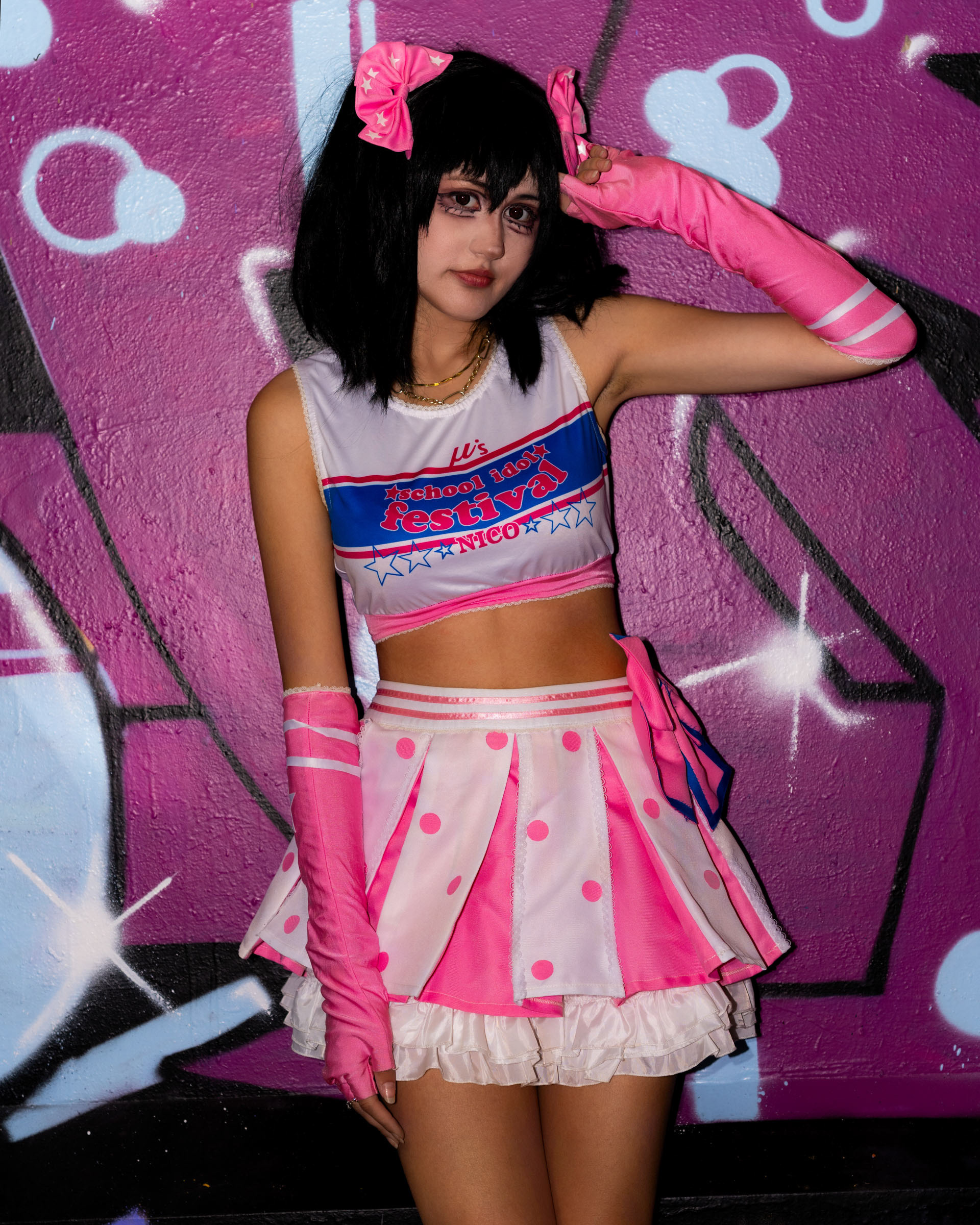 Close-up portrait of may_c0z as Nico Yazawa, giving a peace-sign gesture and showing detailed idol makeup and pink gloves.