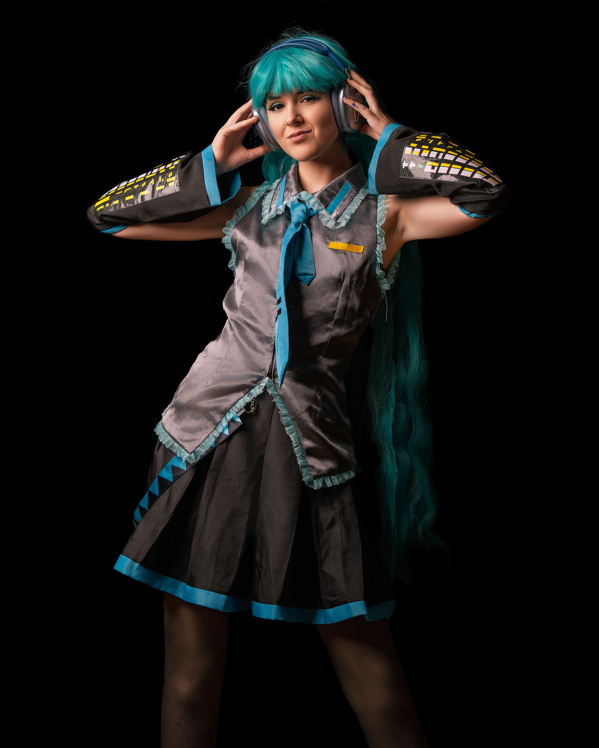 Cosplayer dressed as Hatsune Miku posing calmly in a studio with a dark background, wearing turquoise wig and futuristic headphones.