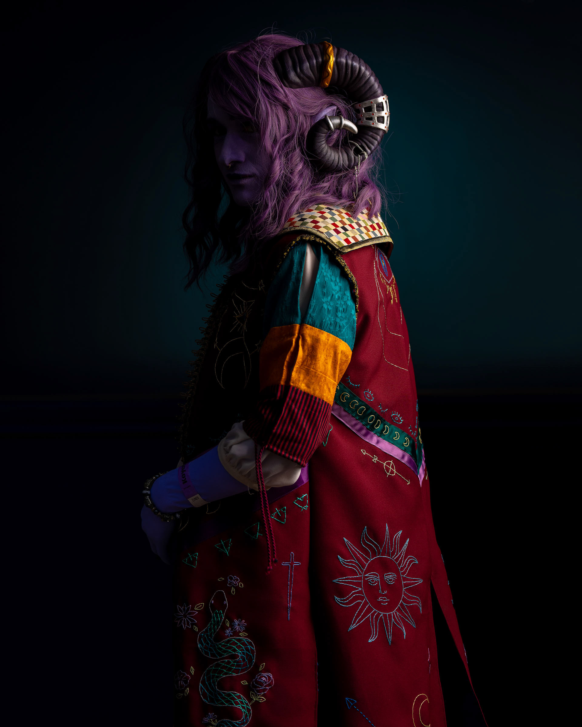 Back-lit Jester Lavorre cosplay showing embroidered red coat with sun and serpent motifs, horns silhouetted against a dark blue background.