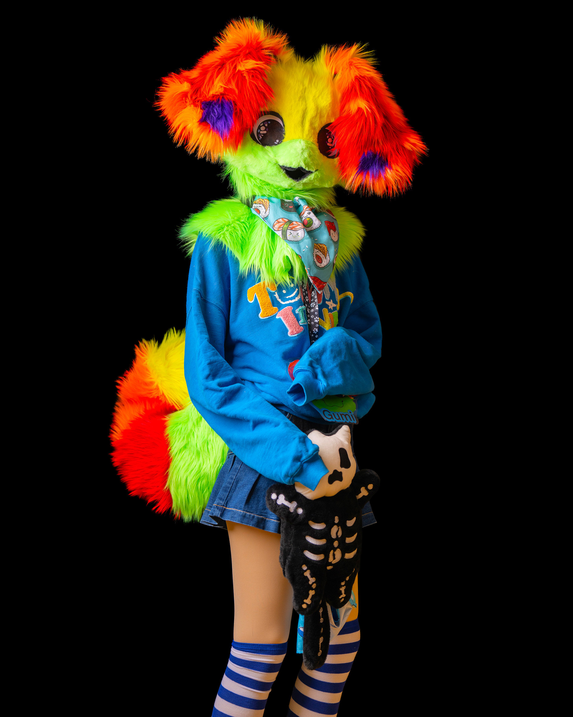 Colorful fursuit cosplay model posing in studio with bright green, orange and yellow costume on black background.