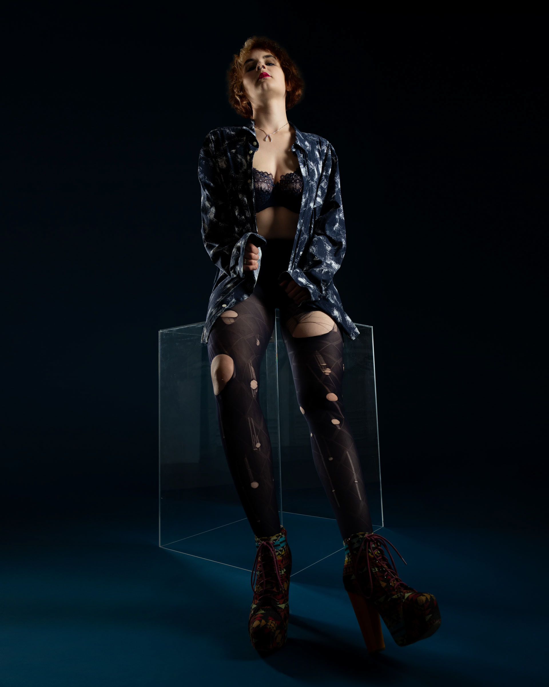 Young female model Sisse posing confidently in lace bra, patterned shirt, and ripped tights while sitting on a glass cube.