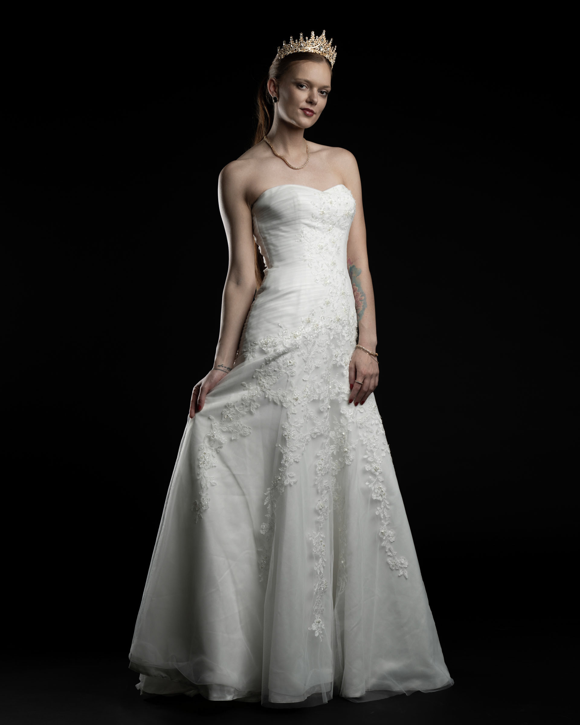Model Noamynte standing in a white bridal gown with a gold crown, posing elegantly in studio lighting.
